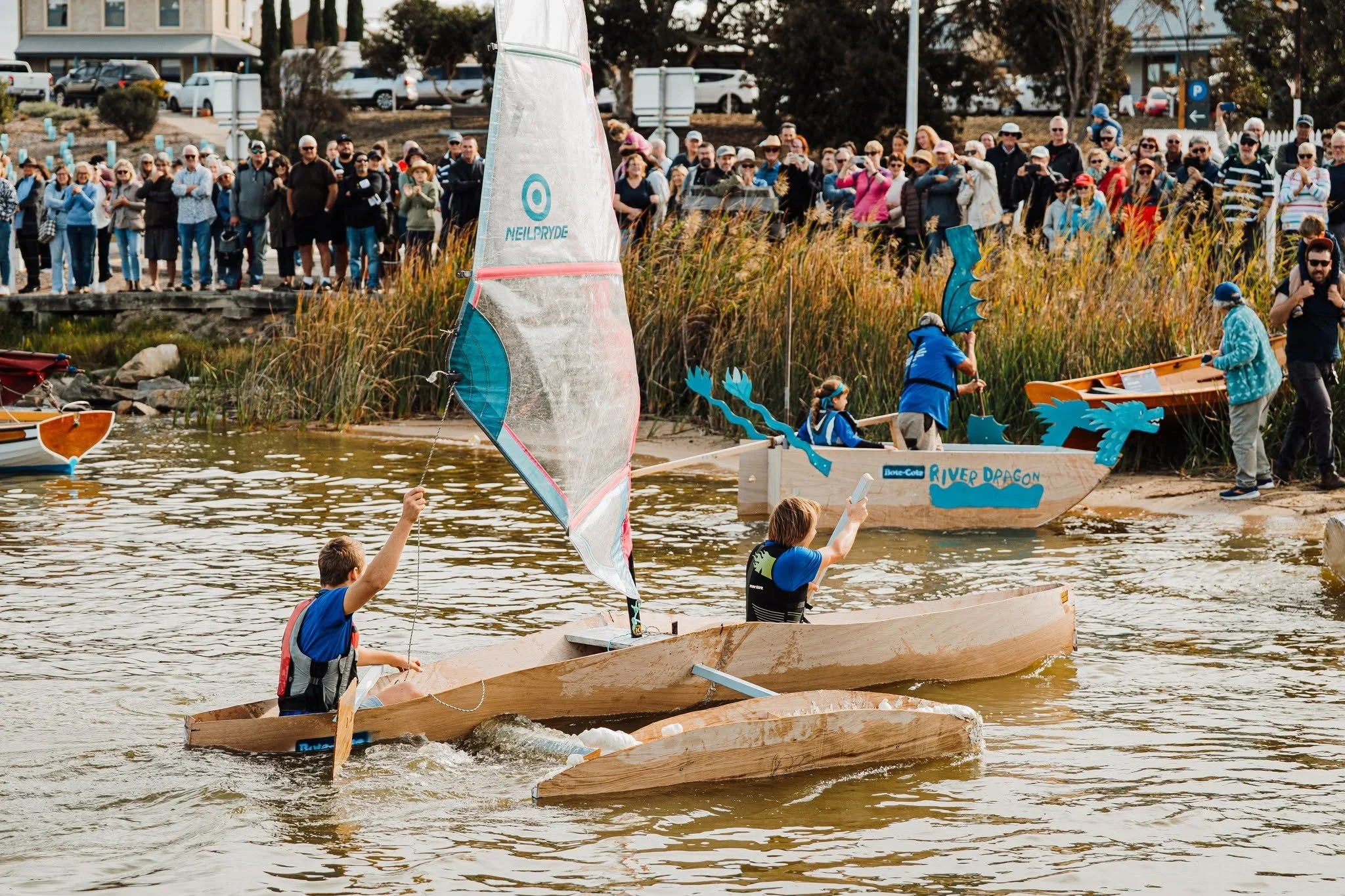 Children participating in a boat race on the water, with a crowd of spectators watching from the shore. Some boats are decorated with dragon designs, and there is a large crowd of onlookers in the background.