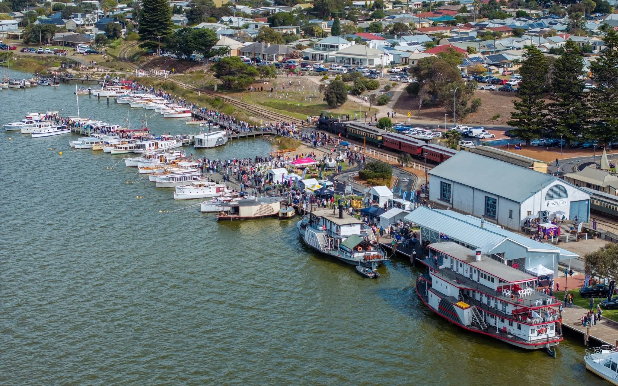 A busy waterfront with numerous boats docked and a crowd of people. There is a train passing through the area and various buildings, including a distillery and a parking lot, with trees and residential houses in the background.