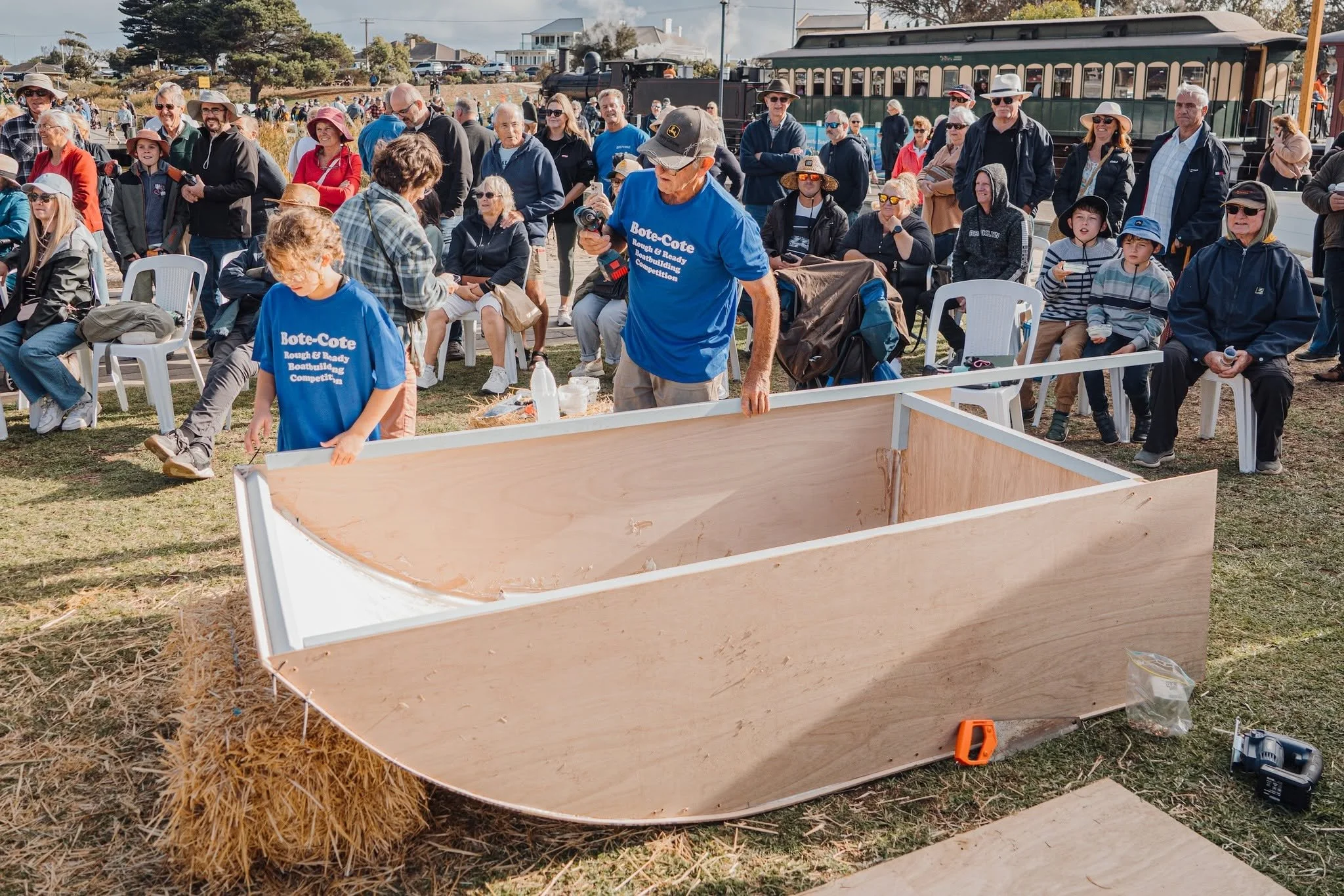 Crowd of people gathered outdoors at a boat-building event, some sitting on chairs, others standing, with a woman in a red jacket and a young girl in a blue t-shirt in the foreground. In the background, a vintage train is visible on a track and resid