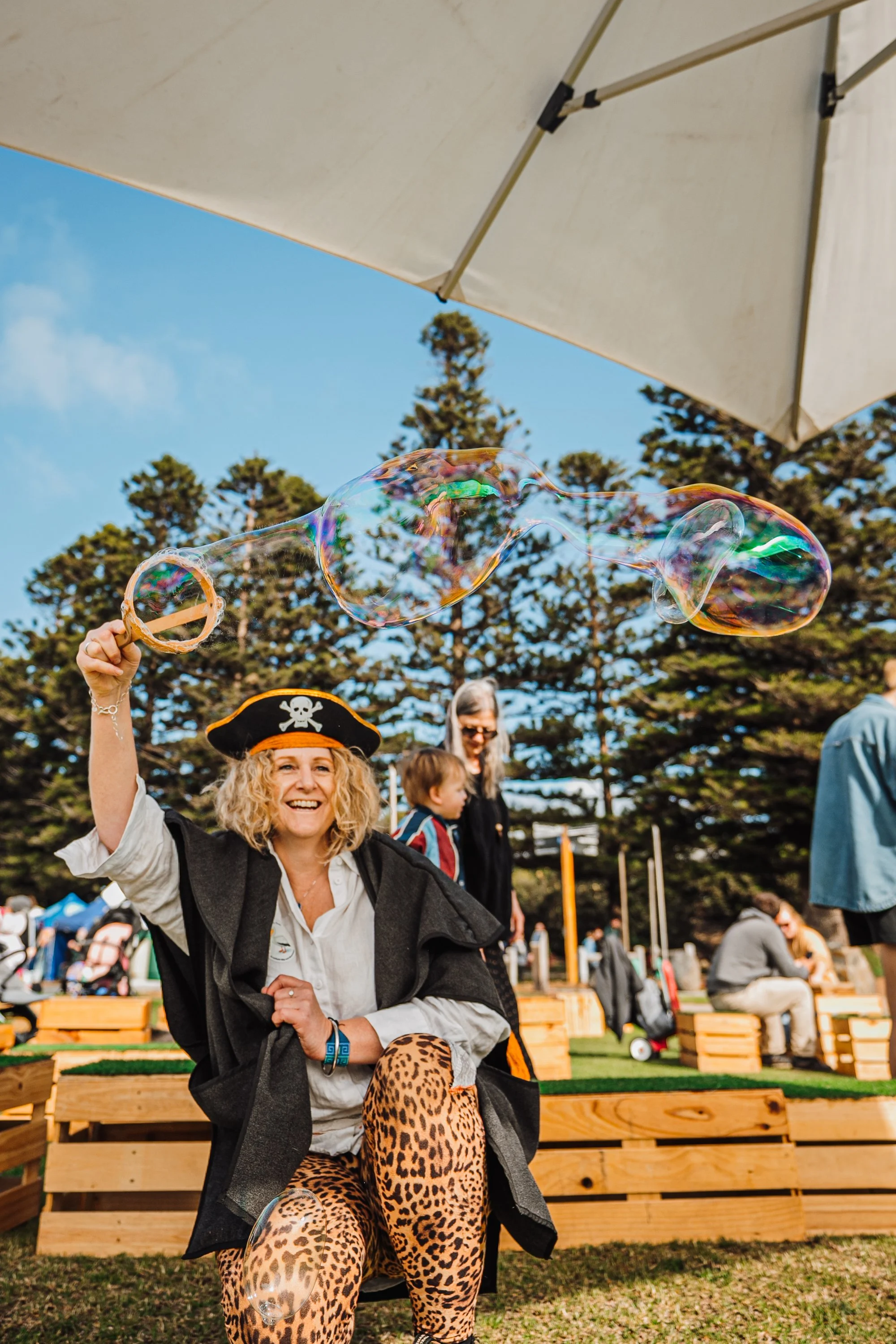 Woman dressed as a pirate with a hat featuring a skull and crossbones, smiling while holding a large soap bubble wand outdoors on a sunny day. There are people in the background and wooden benches in a park setting.
