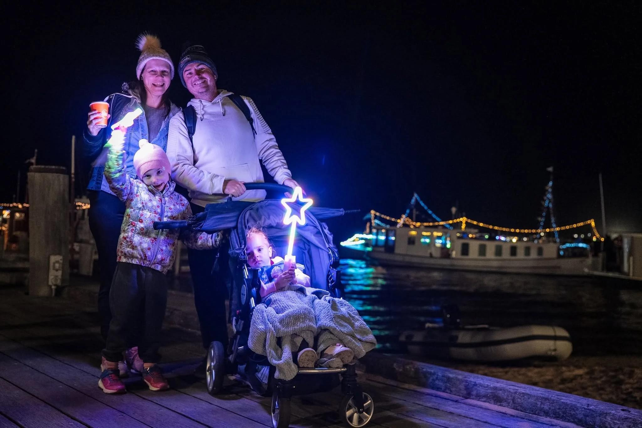 Family of four at night on a dock, with illuminated decorations and a boat in the background. The mother, father, and two young girls are smiling, and one girl is sitting in a stroller holding a glowing star wand.