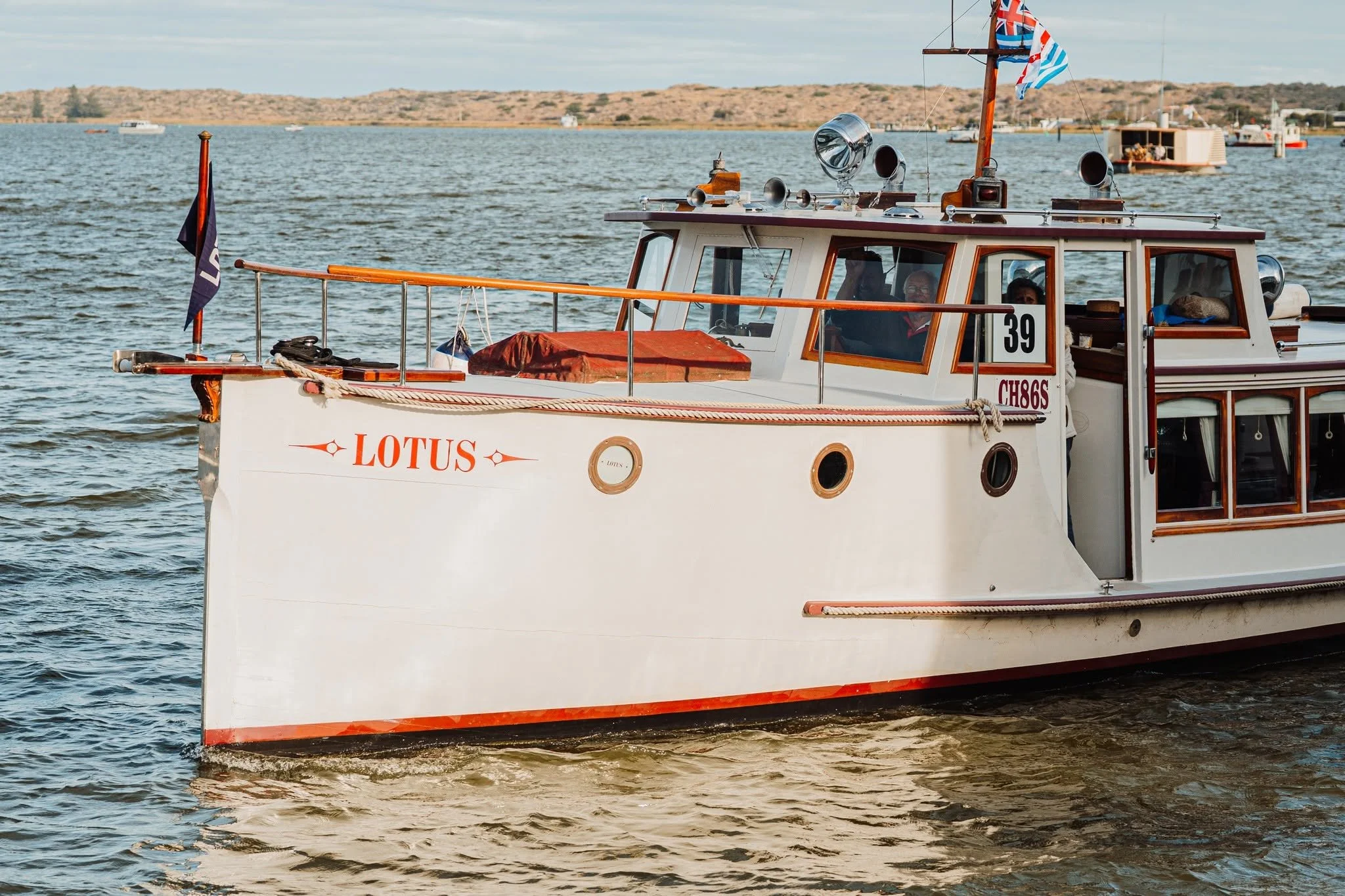 A white boat named Lotus is sailing with two people inside. The boat is decorated with flags, and there are various equipment and accessories on the roof and deck. In the background, there are other boats and a shoreline with sandy hills.