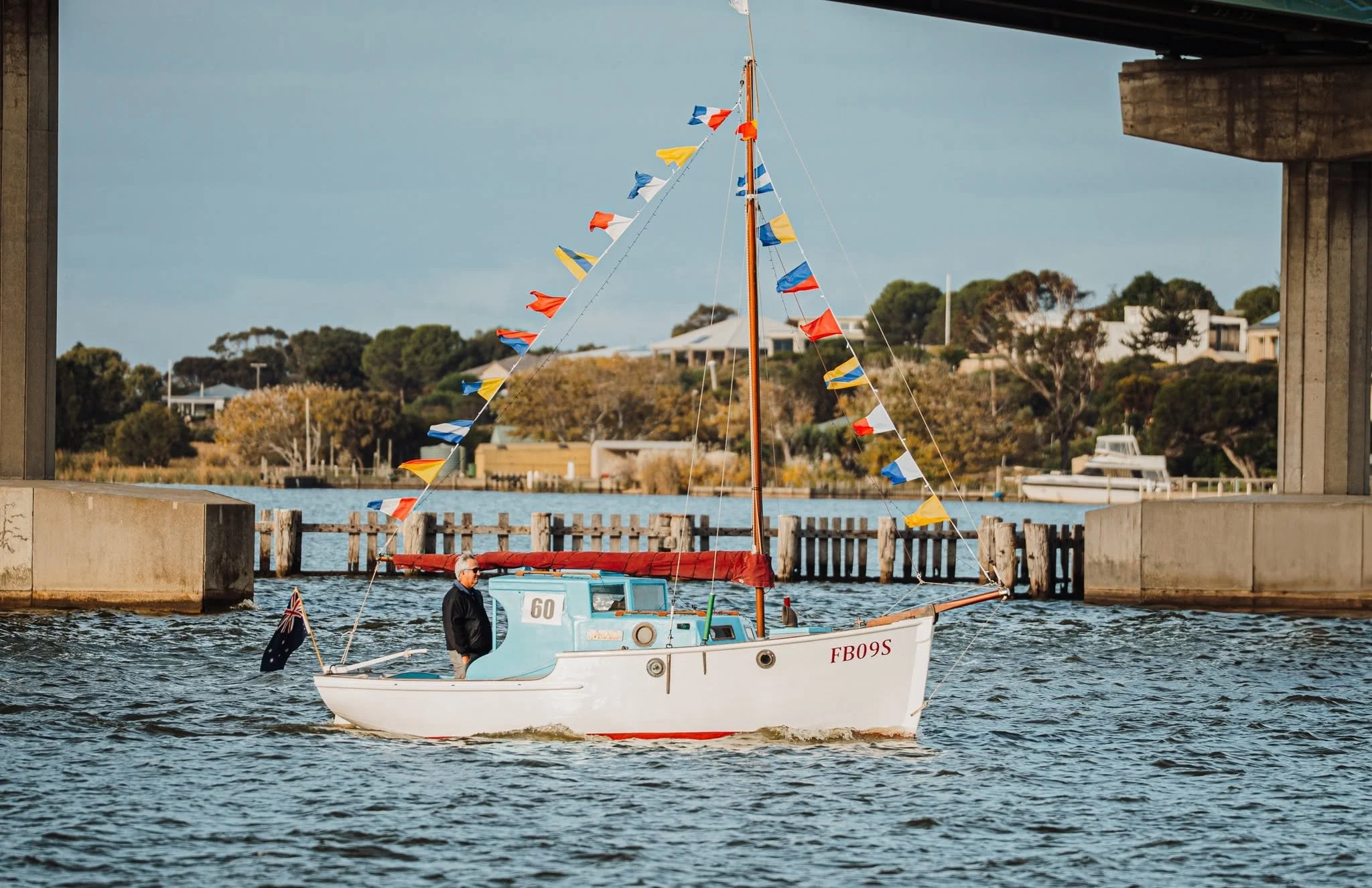 A small white sailboat with colorful flags on its mast, sailing under a bridge in a waterway with houses and trees on the shoreline.