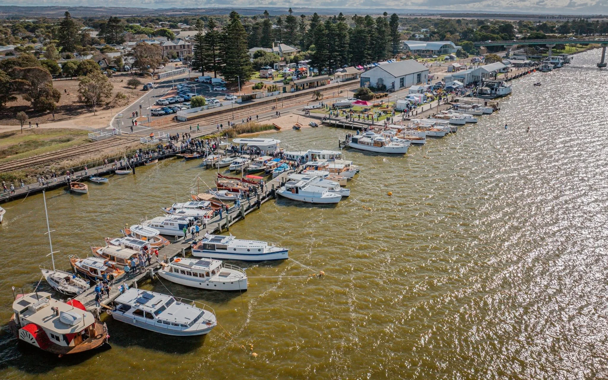 Aerial view of Goolwa Wharf with numerous boats docked and a bustling waterfront area with tents and people, next to a river with a bridge in the background.