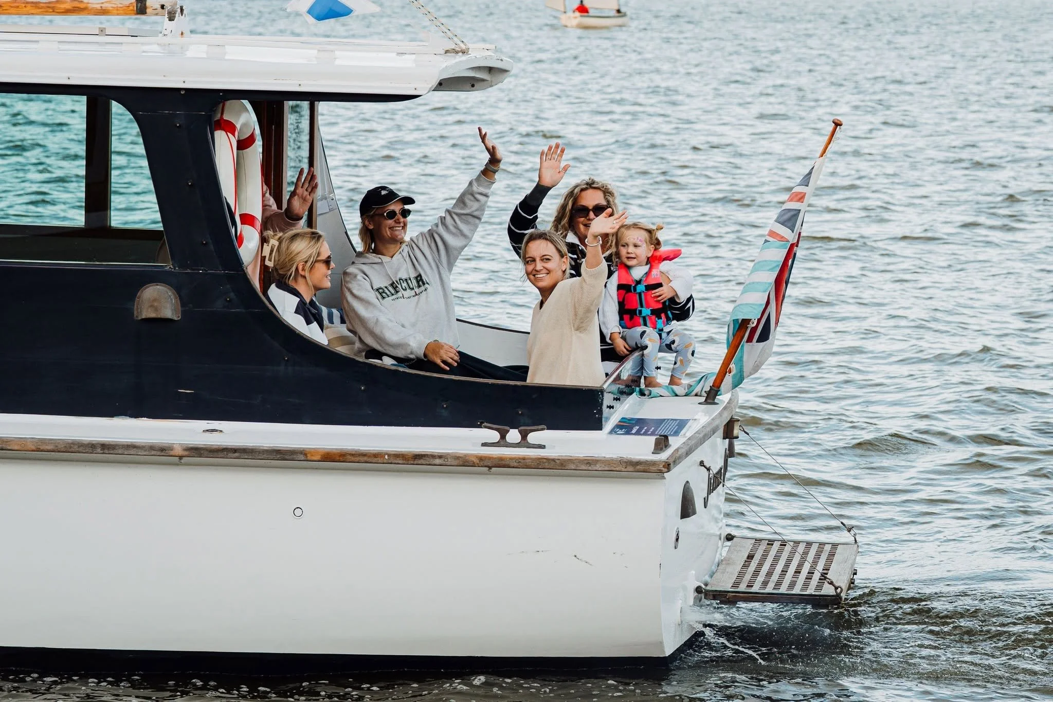 A group of five people, including a child, on a boat waving and smiling at the camera, with water and other boats in the background.