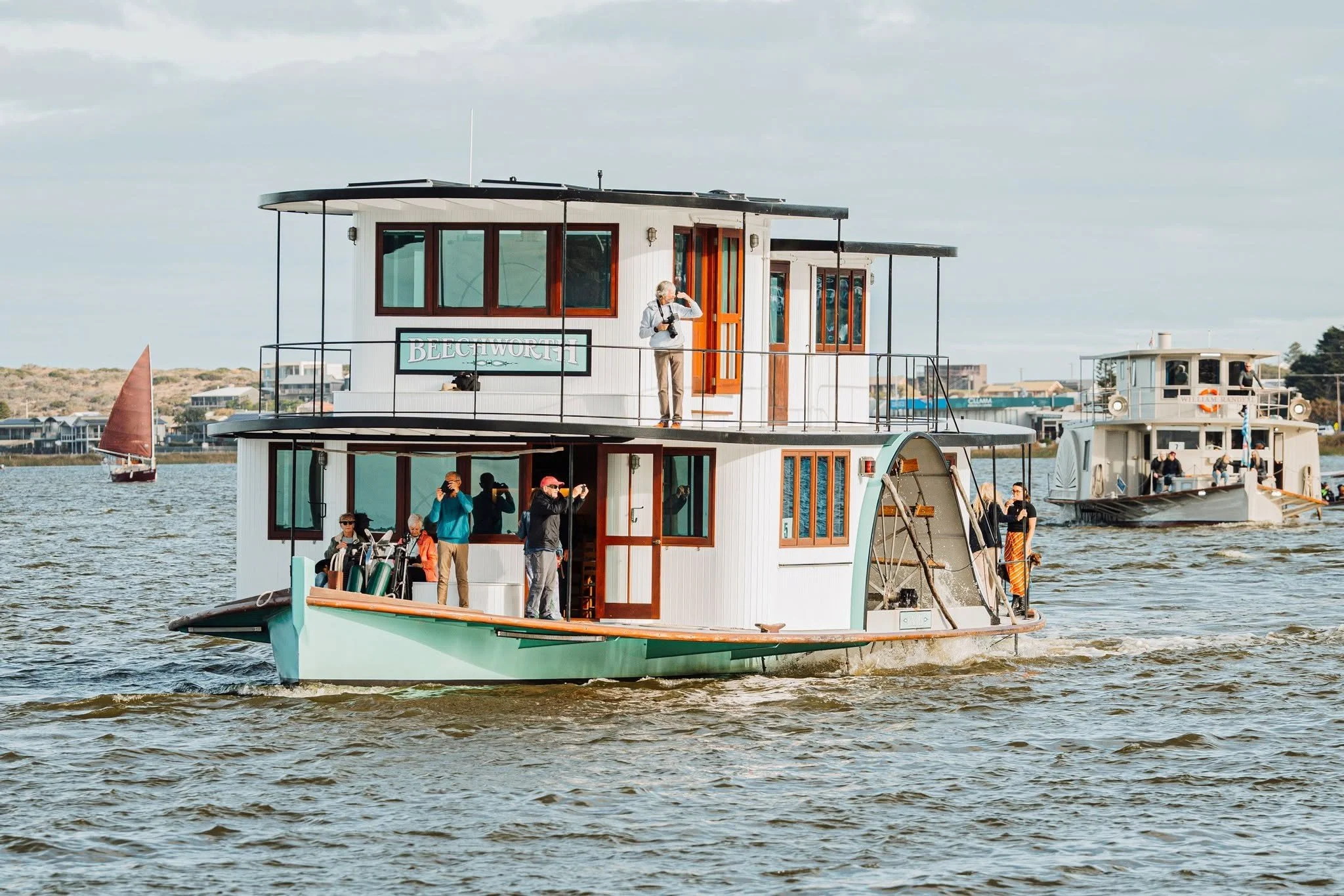 A white paddle steamer with multiple windows and a balcony on the river, with people onboard taking photos and a few boats and houses in the background under a cloudy sky.