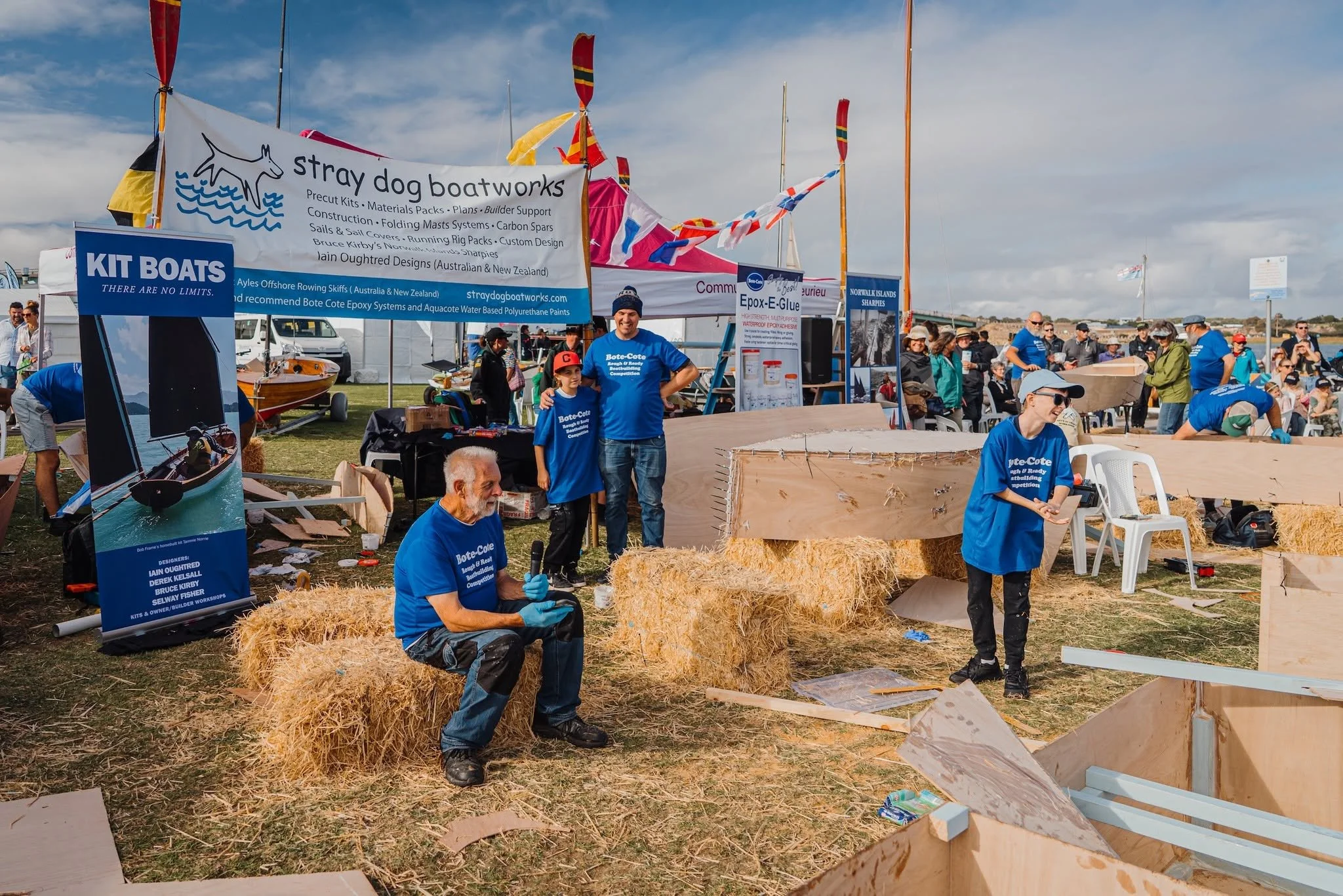 People at a booth, building wooden boats and displaying banners related to stray dog boatworks and kayak designs, with some hay bales and equipment visible.
