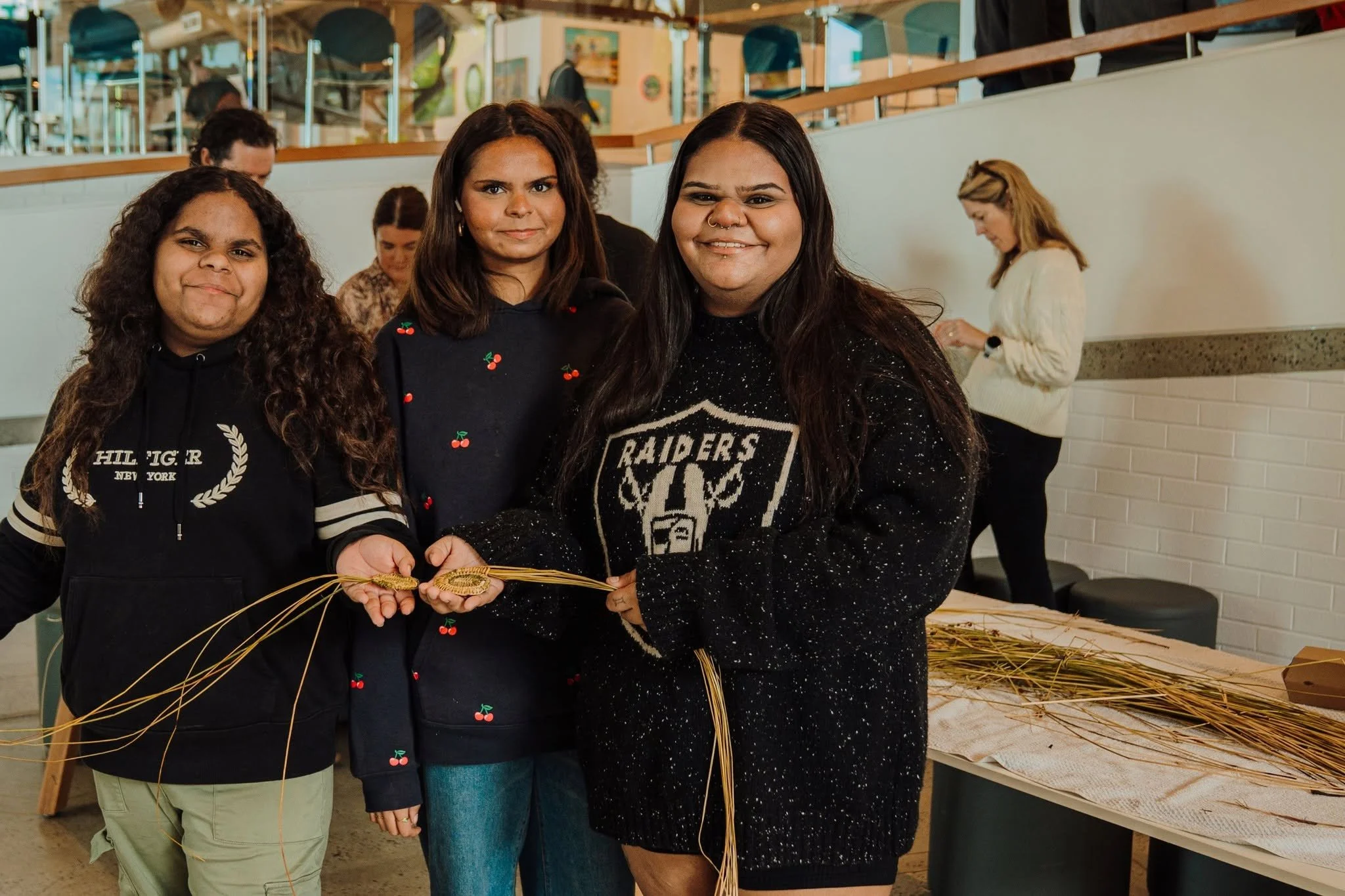 Three women standing close together in a room, holding straw decorations, with a group of people in the background. The women are smiling and appear to be participating in a craft activity.