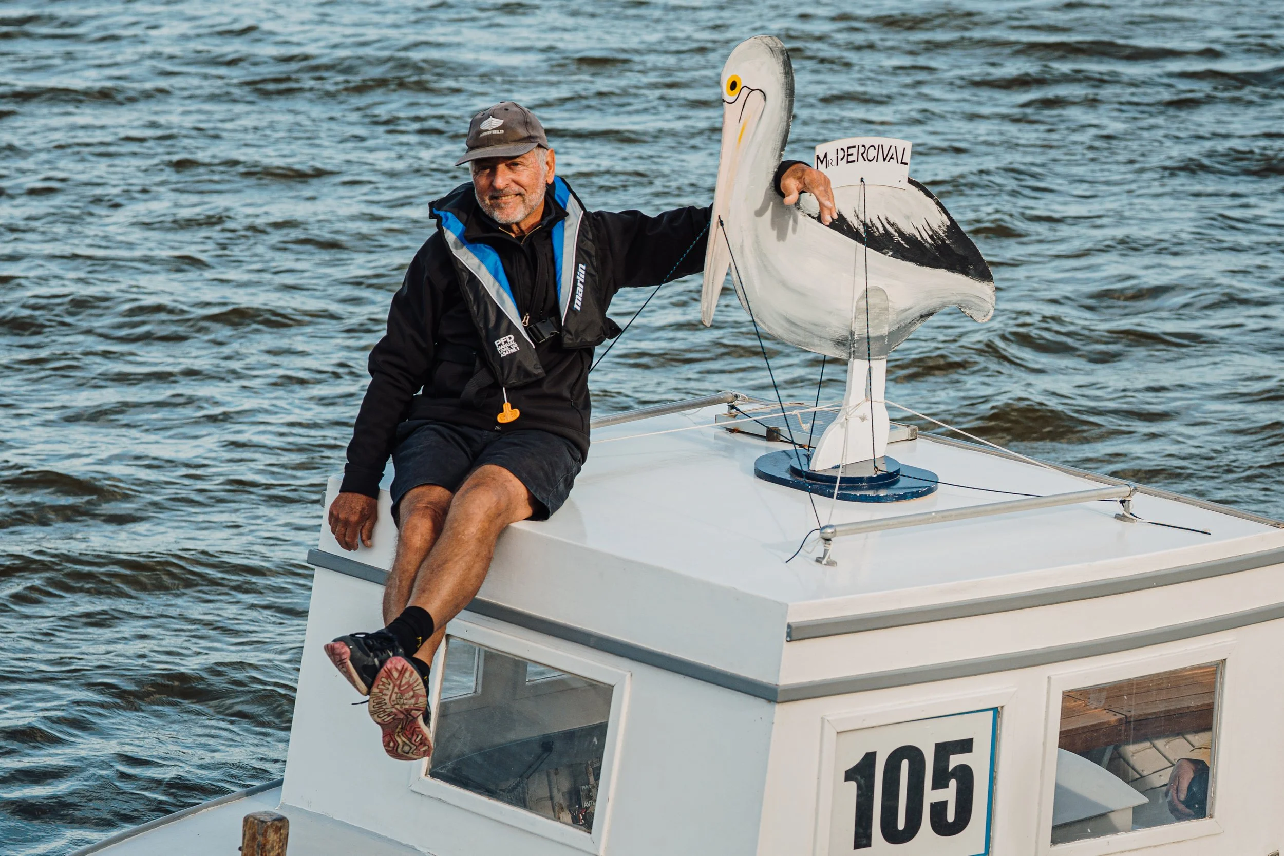 A boatie sitting on the edge of his boat with his legs dangling over the side, smiling, and holding a decorative painted pelican.