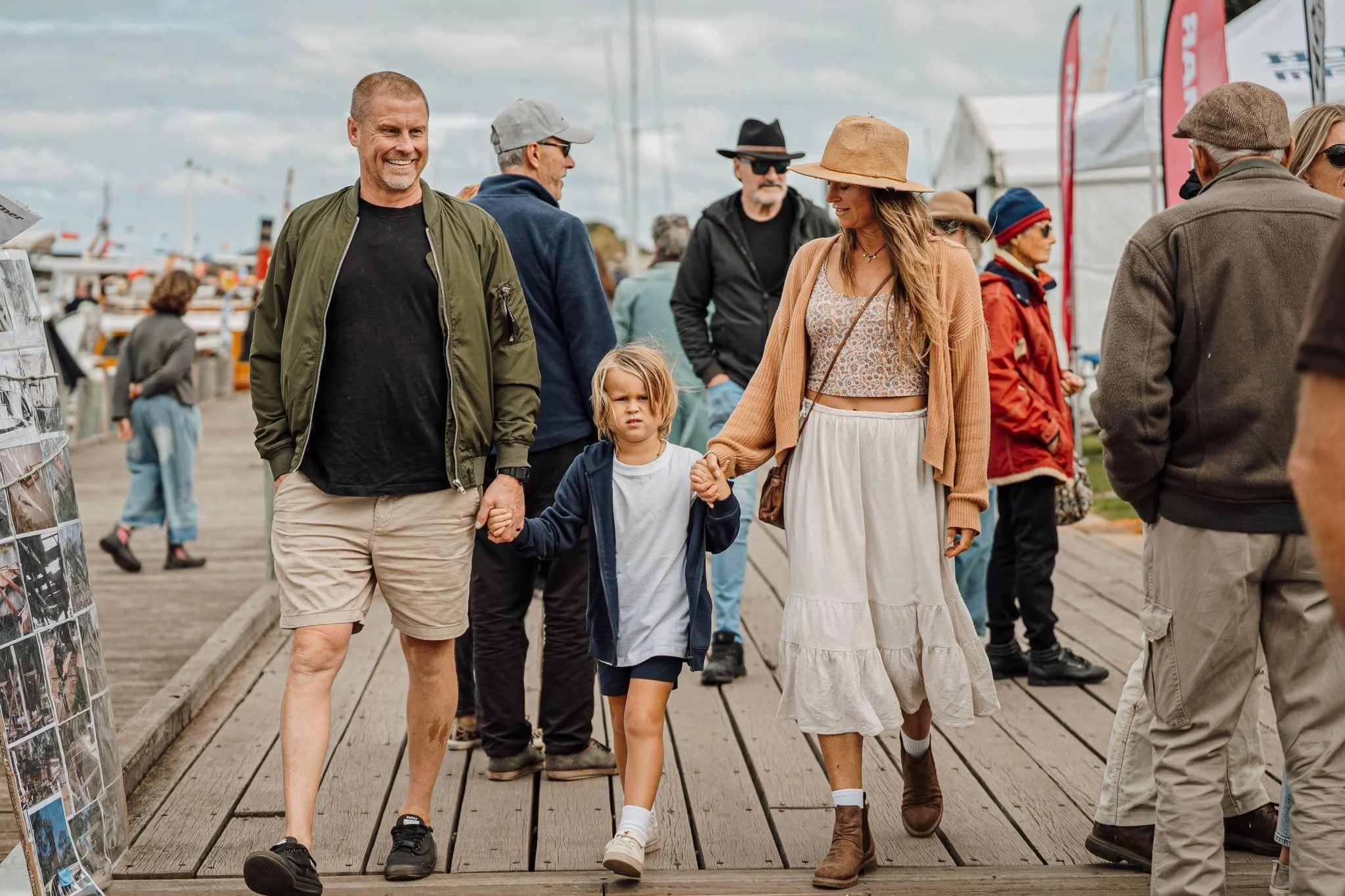 Group of people walking and talking on a wooden pier near boats and tents, some wearing hats and casual clothes, with a photo display stand on the side.