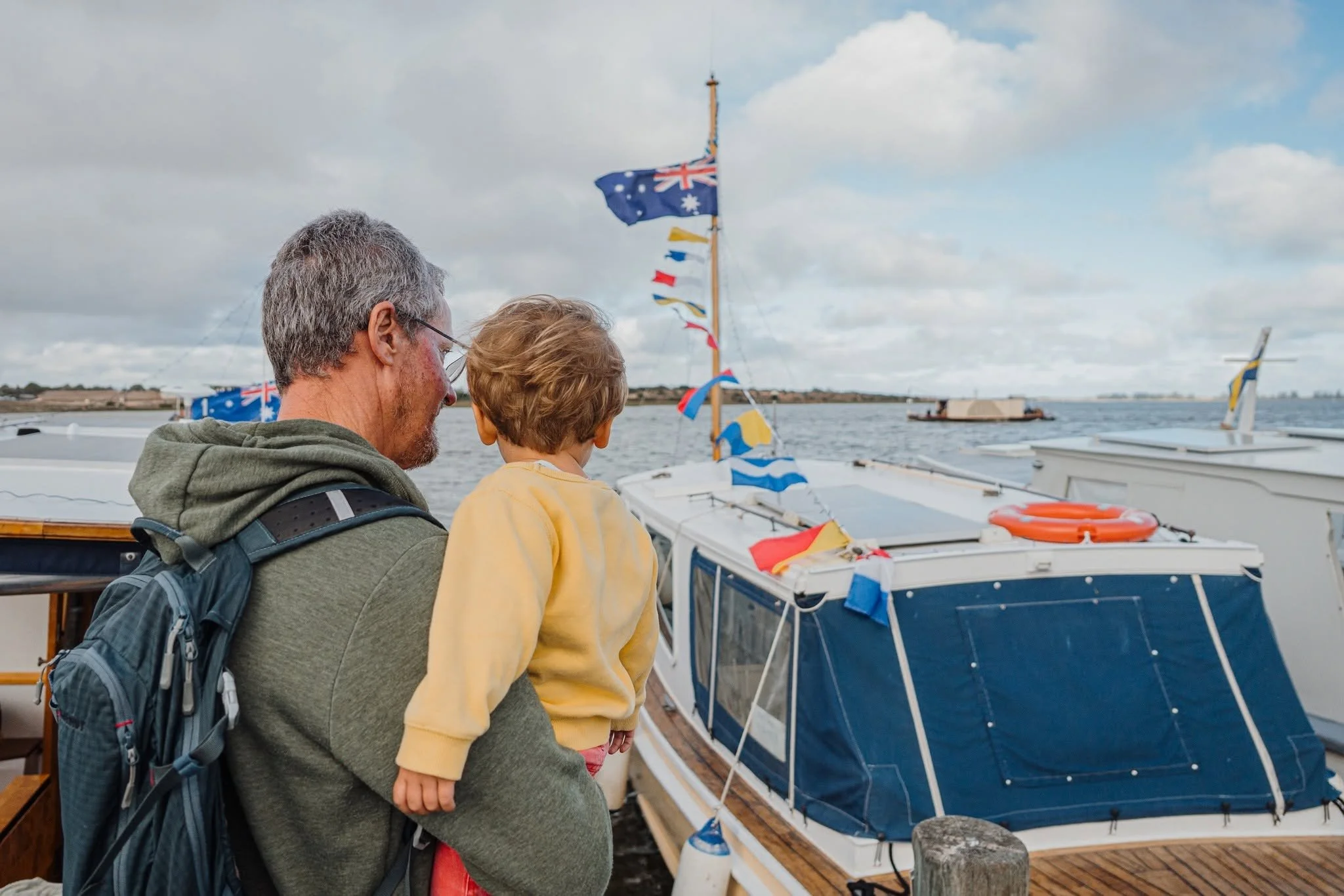 A man with gray hair and glasses carrying a backpack, holding a young child in a yellow jacket, on a boat dock looking at a sailboat with Australian flags, water, and cloudy sky in the background.