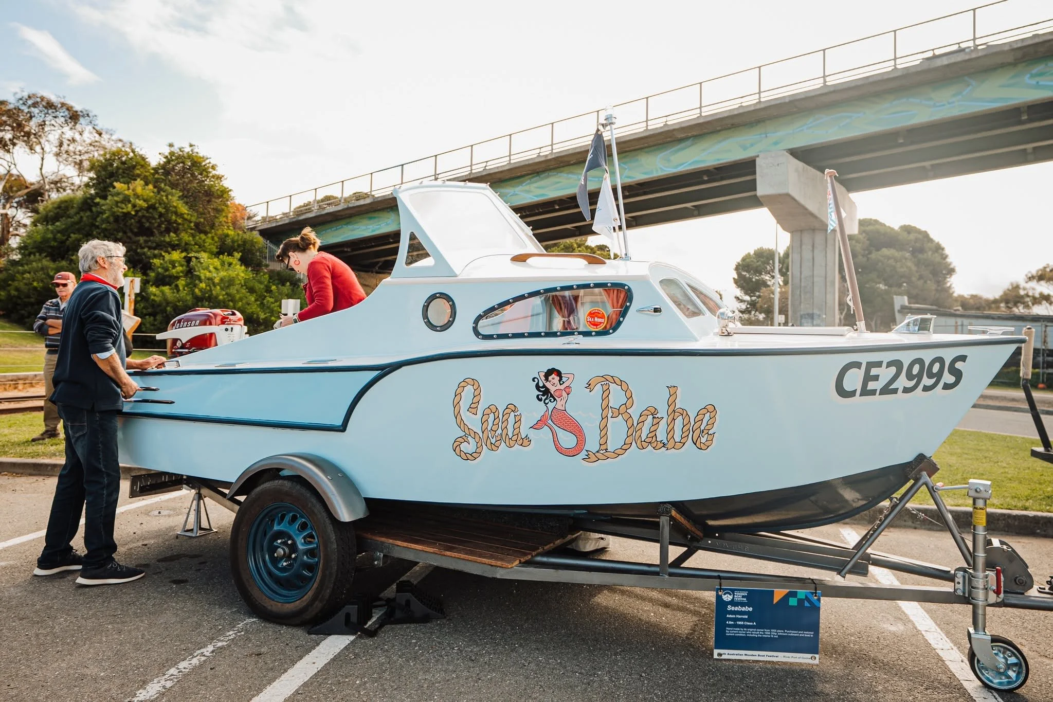 A small sailboat on a trailer, decorated with pin-up style artwork of a mermaid and nautical ropes, with the words "Sea Babe" painted on the side. Several people are examining the boat outdoors near a bridge.