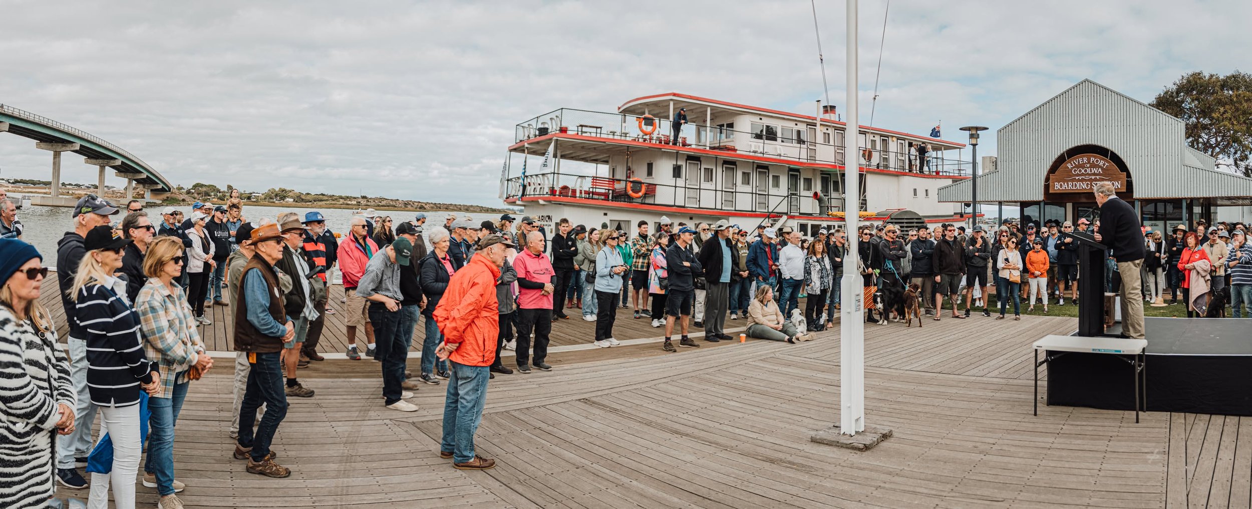 A crowd of people gathered in front of a docked river cruise boat at Goolwa Wharf. The scene is outdoors under a cloudy sky.