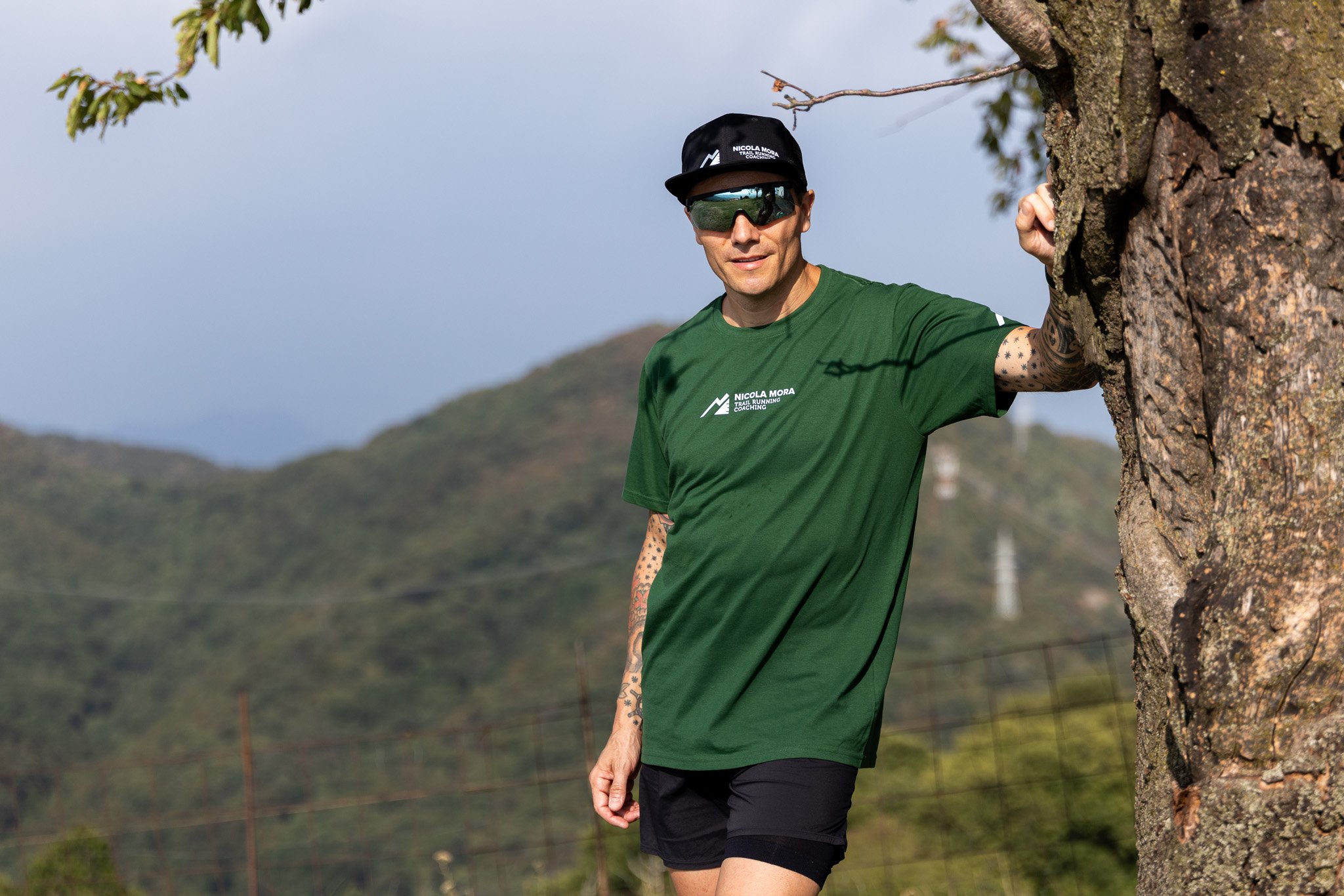 Un uomo corre all'aperto con abbigliamento da corsa, indossa occhiali da sole e cappellino, si appoggia a un albero in un paesaggio naturale con montagne e cielo sereno.