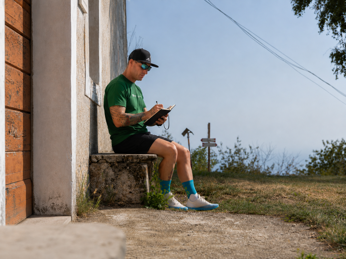 Uomo seduto su un muretto di pietra, che scrive su un taccuino, indossa una maglietta verde, pantaloncini neri, calze blu con motivi, scarpe bianche, cappellino nero e occhiali da sole, all'aperto in un giorno soleggiato.