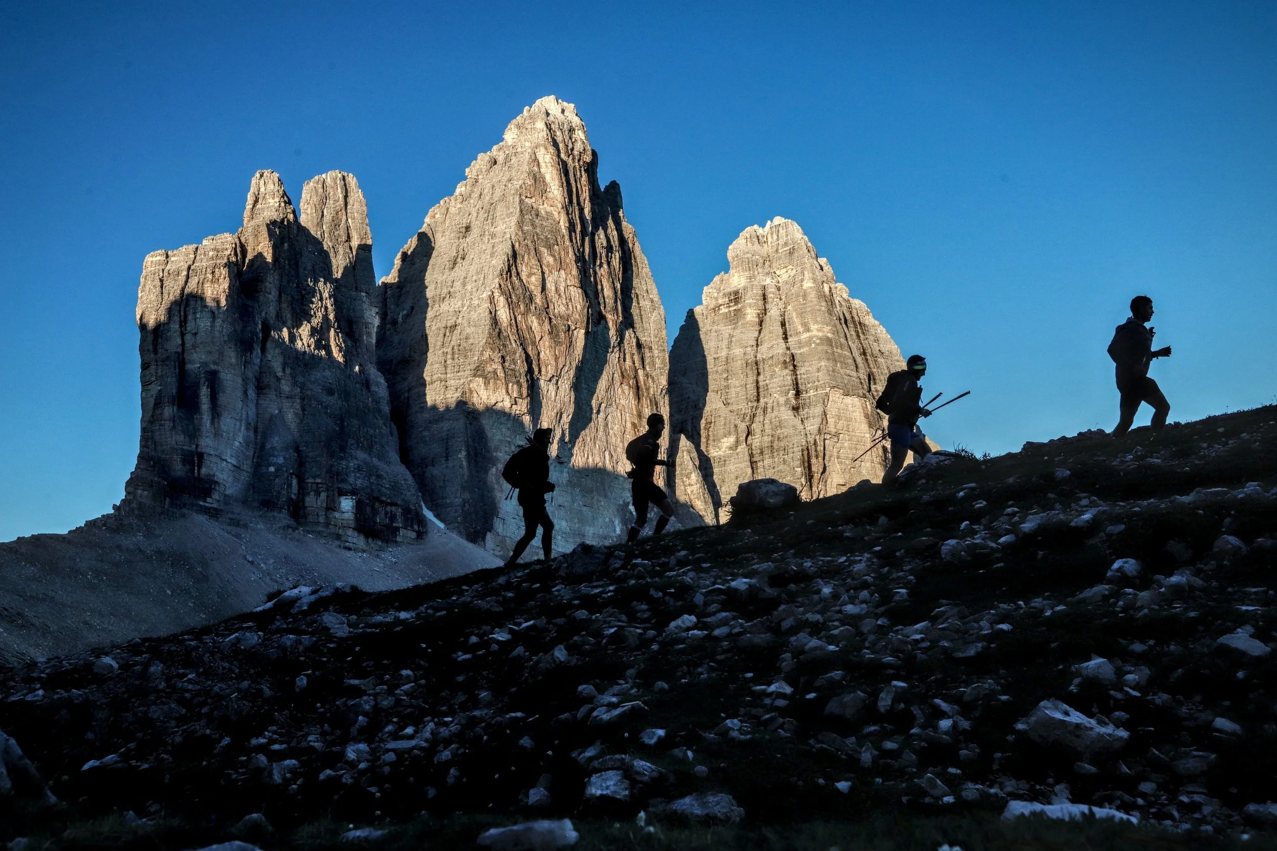 Tre escursionisti camminano in un paesaggio montano con delle alte pareti rocciose e un cielo azzurro sullo sfondo.
