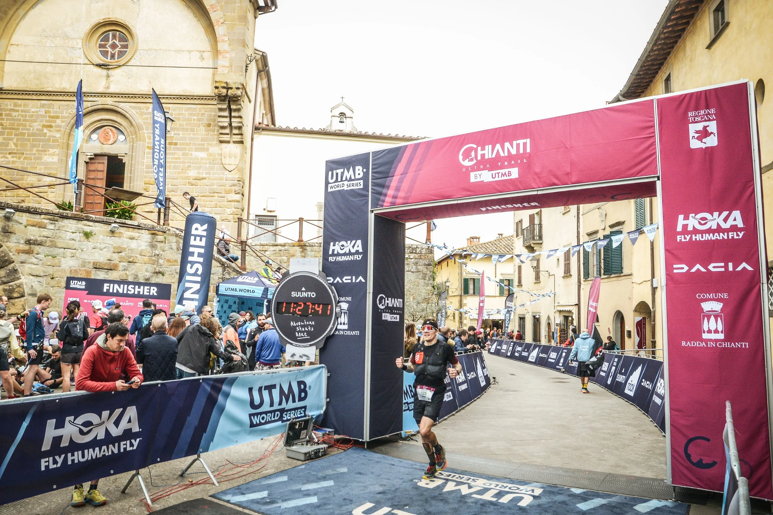 Finisher che attraversa il traguardo di una corsa ultratrail in un centro storico con edificio in pietra, sotto un arco con bandiere e cartelli sponsorizzati.