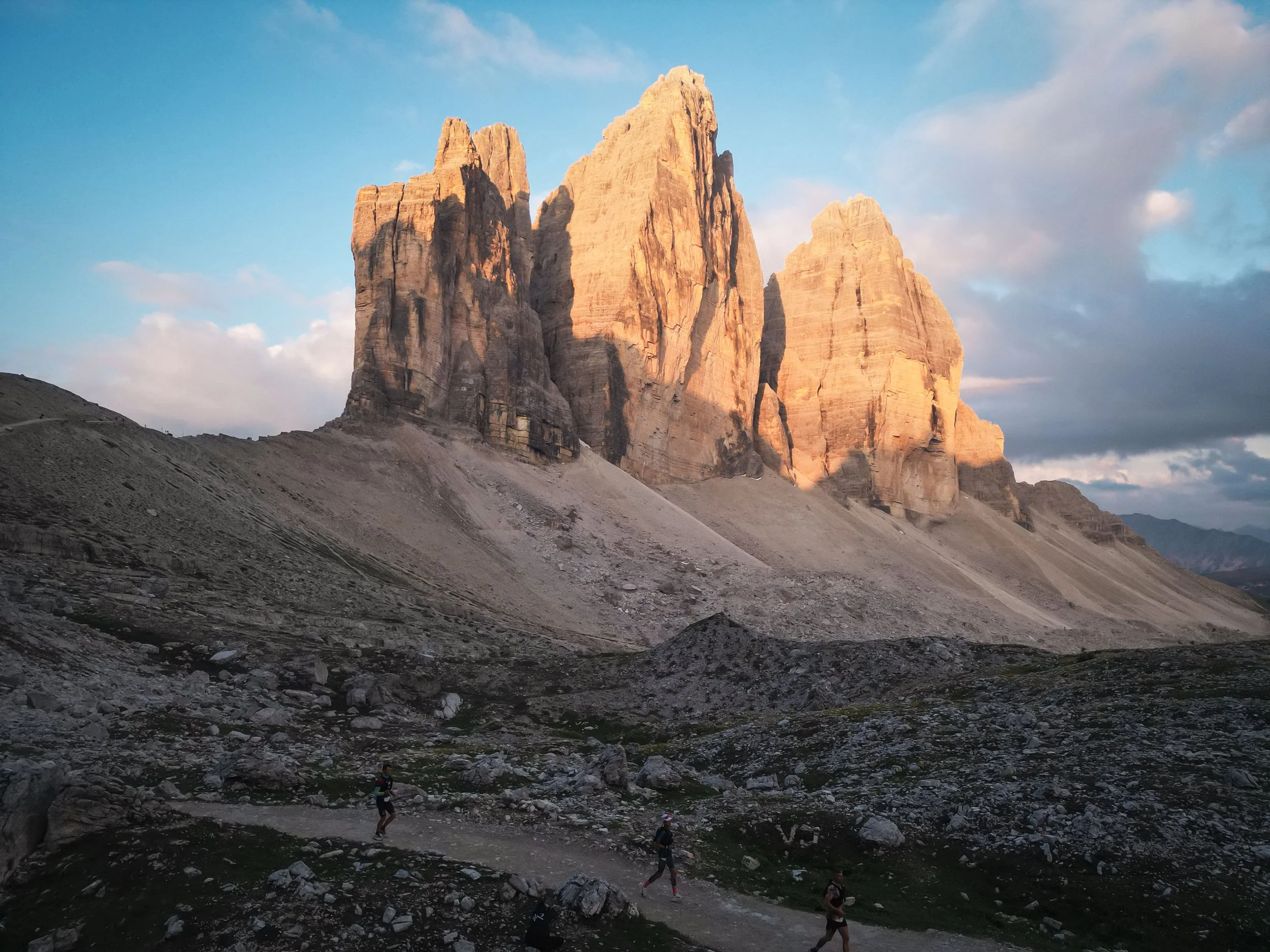 Immagine di imponenti pareti rocciose di una montagna o scogliera, con un cielo azzurro e alcune nuvole, e alcuni escursionisti che camminano su un terreno roccioso in primo piano.