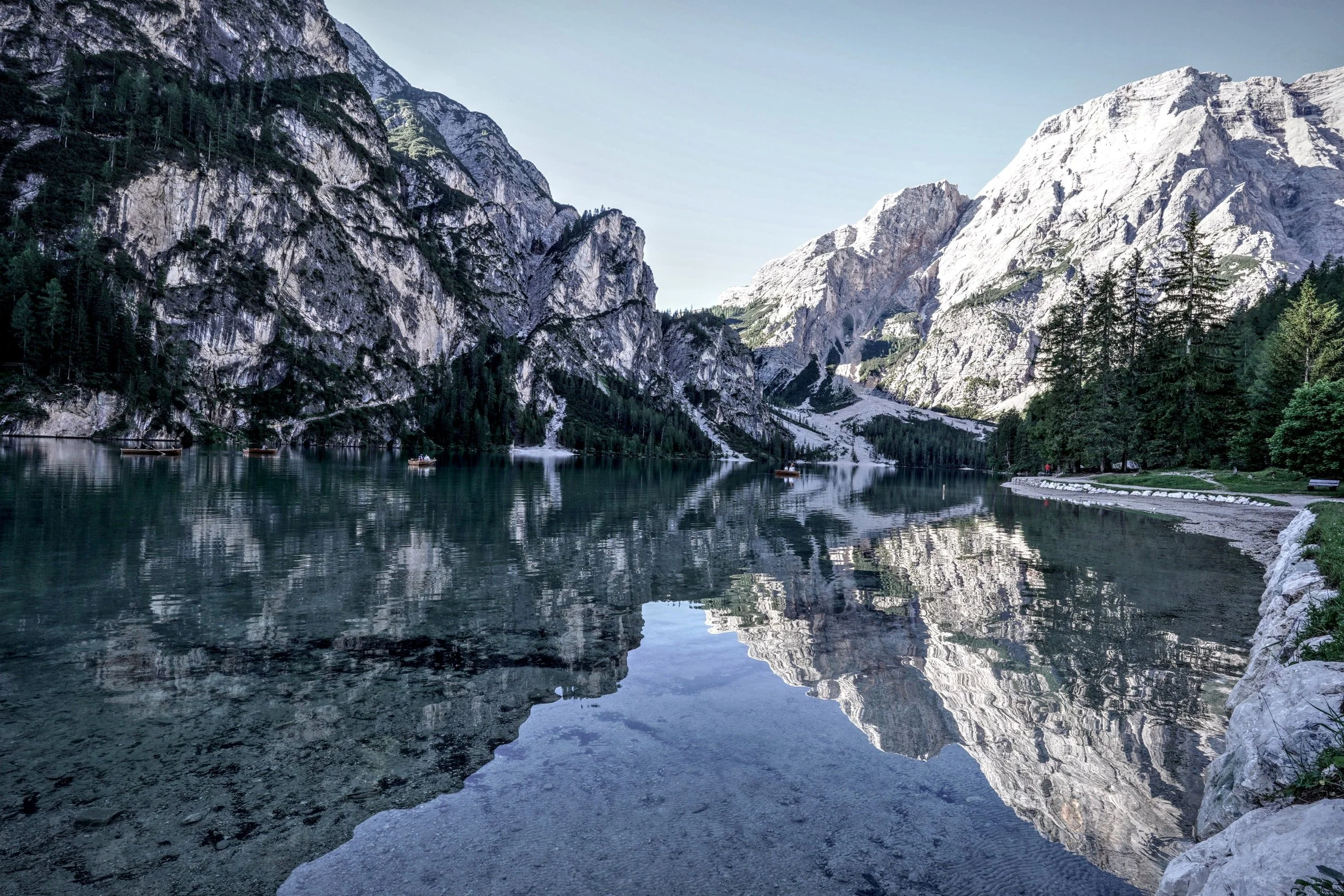 Un lago meraviglioso circondato da alte montagne con pareti rocciose e pini, con alcune barche visibili sull'acqua e una passeggiata lungo la riva.