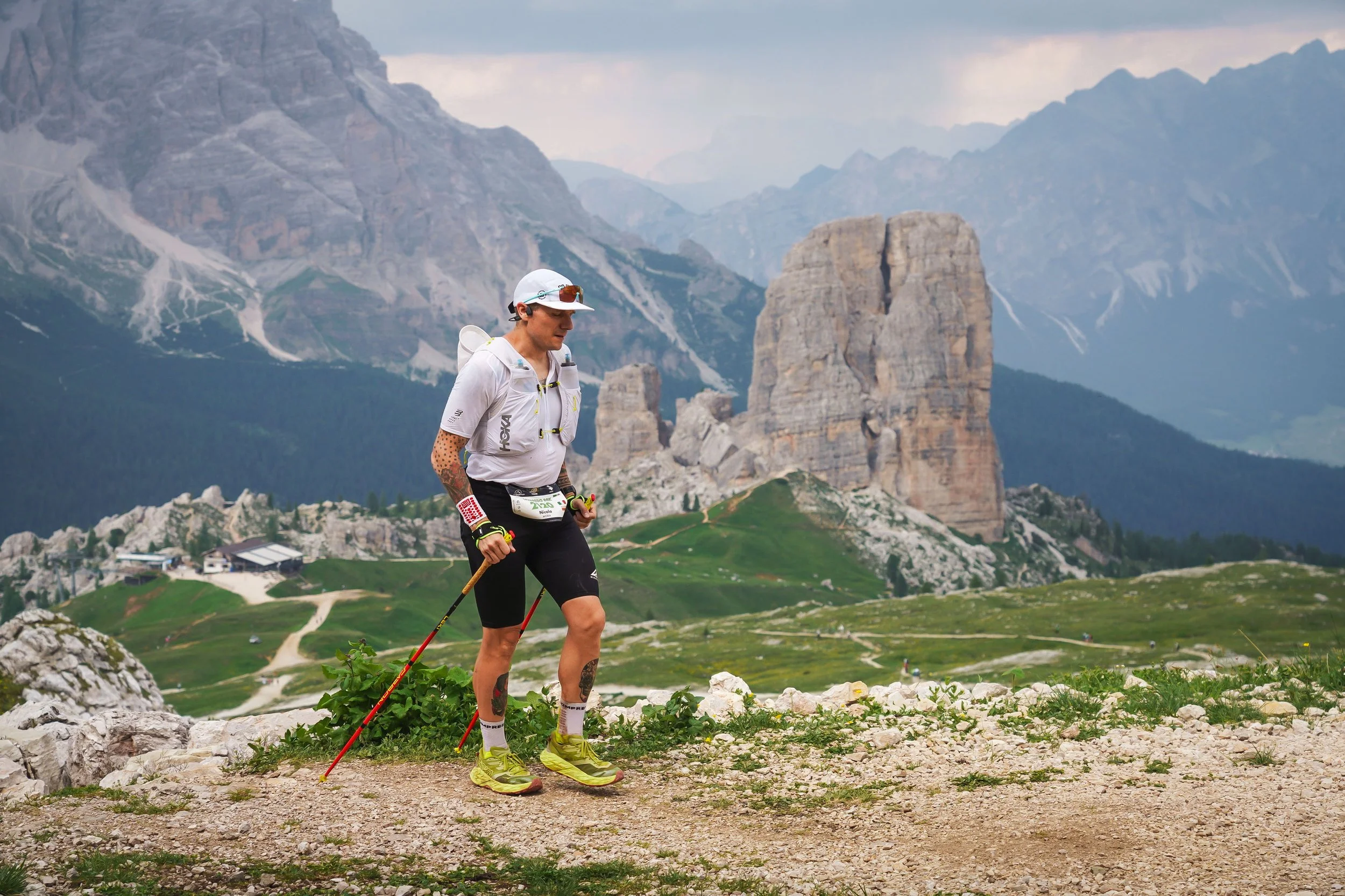 Uomo che pratica nordic walking nelle montagne con rocce e verdi pascoli, sfruttando bastoncini e abbigliamento sportivo, con un paesaggio montano sullo sfondo.