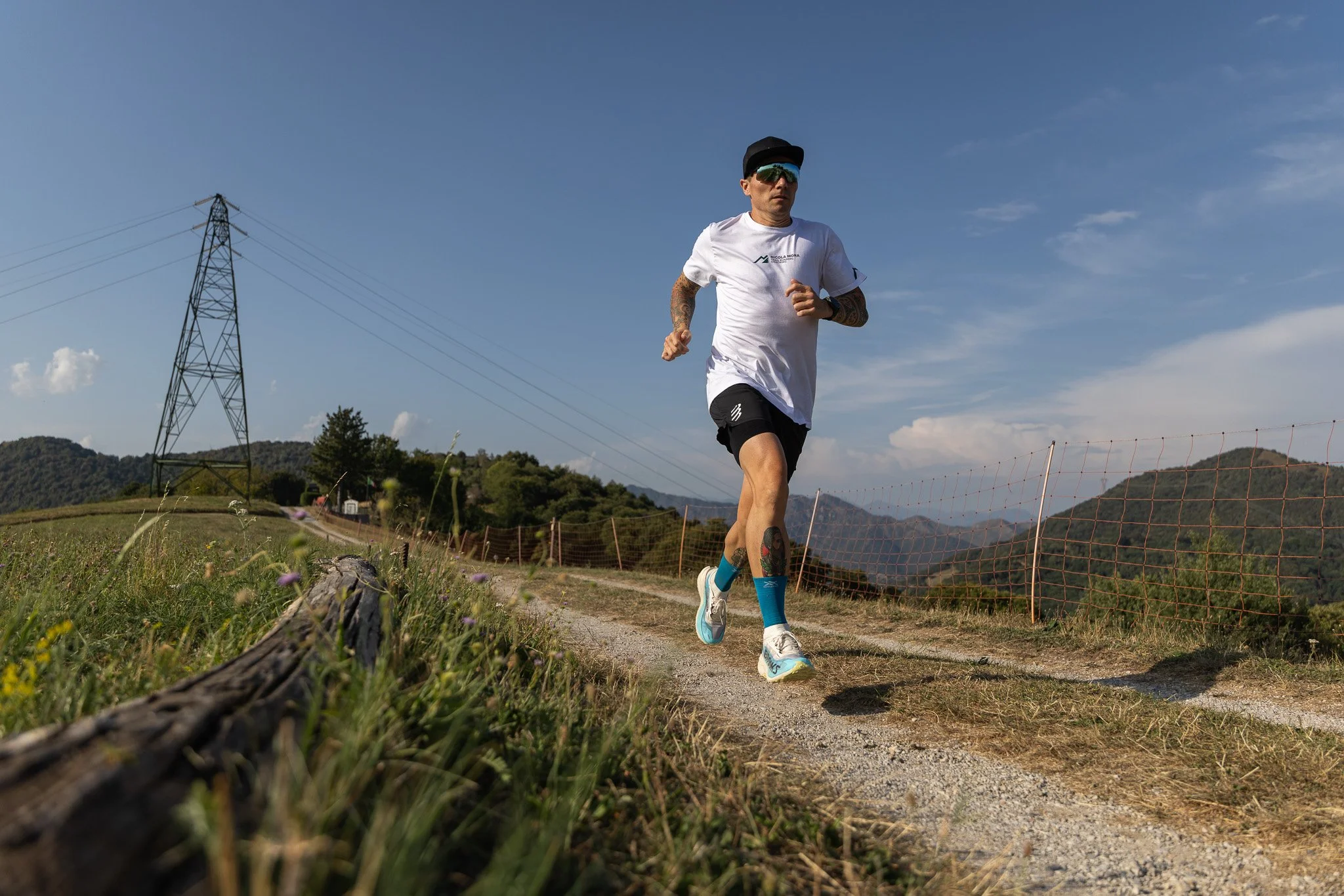 Uomo che corre lungo un sentiero di campagna, vestito sportivamente, con uno sfondo di cielo azzurro e colline verdi, vicino a un'alta torre di trasmissione elettrica.