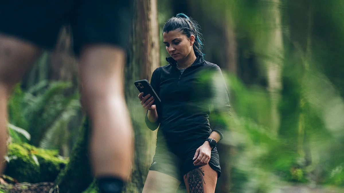 Una donna che si trova in un bosco, che indossa abbigliamento sportivo nero e guarda il telefono, con un paio di gambe (probabilmente di un'altra persona) in primo piano.