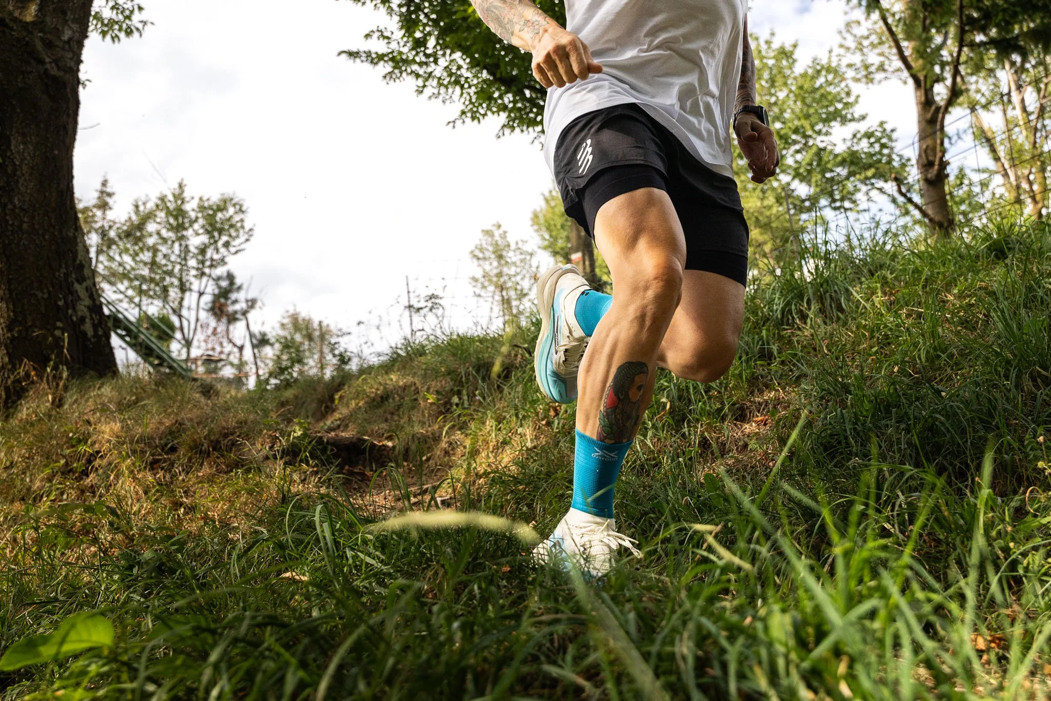 Uomo che corre lungo un sentiero immerso nella natura, con abbigliamento sportivo e tatuaggi visibili sulle gambe e le braccia.