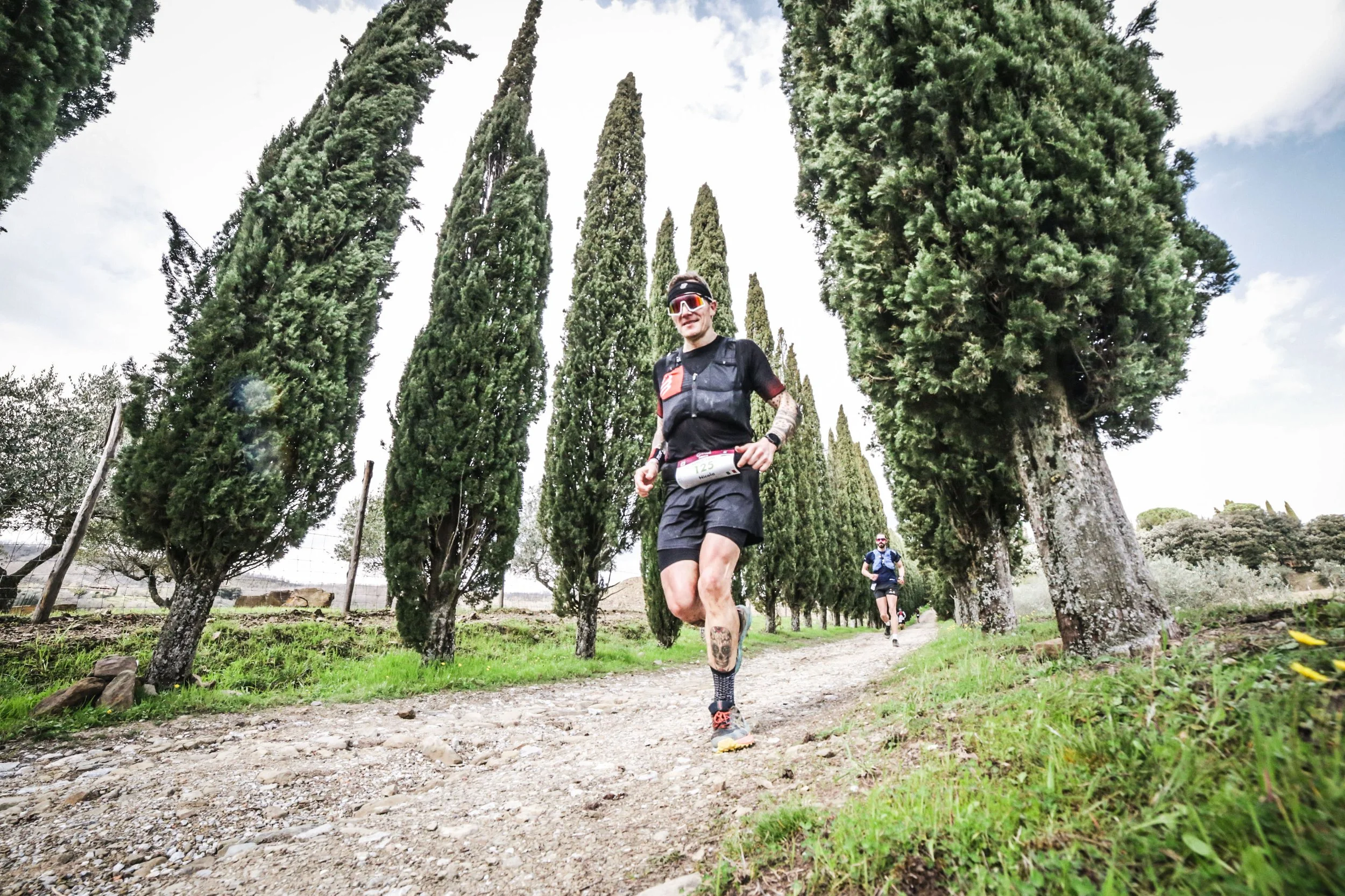 Uomo che corre su un sentiero di campagna circondato da alberi alti di cipresso, con altri runner alle sue spalle.