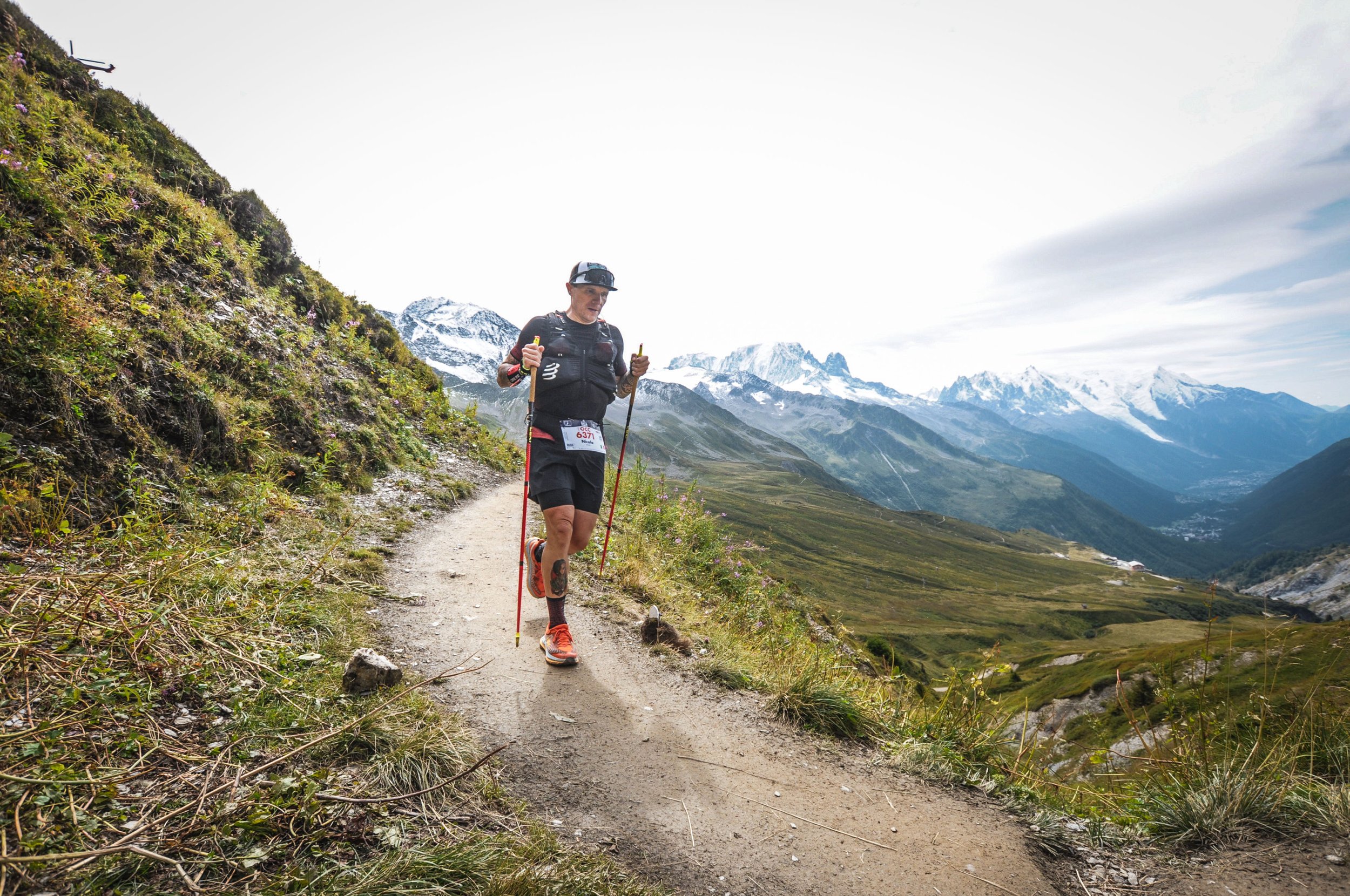 Uomo che corre in montagna con scarpe da trail running, cappellino e bastoncini lungo un sentiero in un paesaggio alpino con montagne innevate sullo sfondo