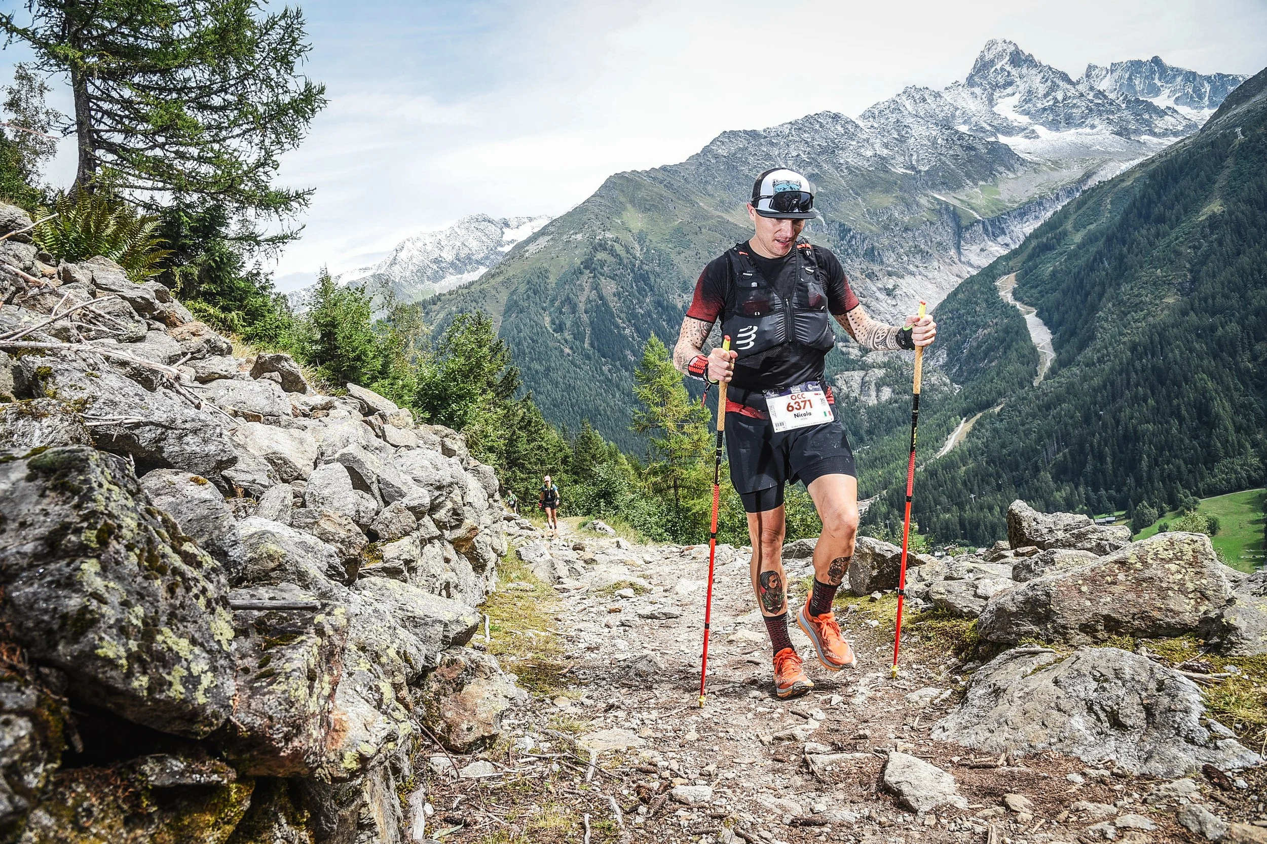 Uomo che fa trail running su un sentiero di montagna, circondato da paesaggi di montagne con neve e foreste, indossando abbigliamento sportivo e attrezzature per l'outdoor.