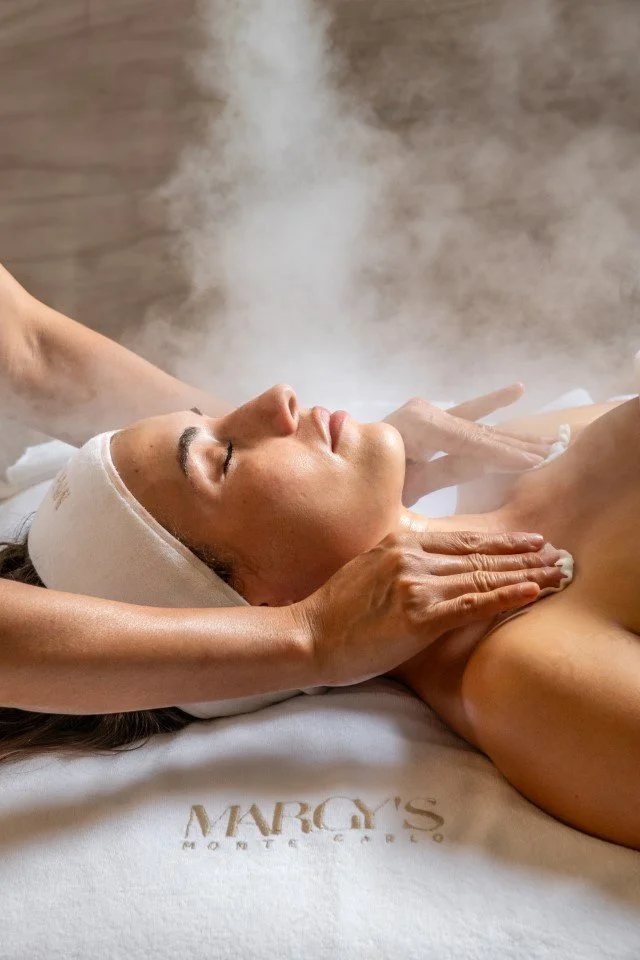 Therapist's hands cradling a client's face during a facial massage, with the Margy's Monte Carlo logo visible at the bottom.