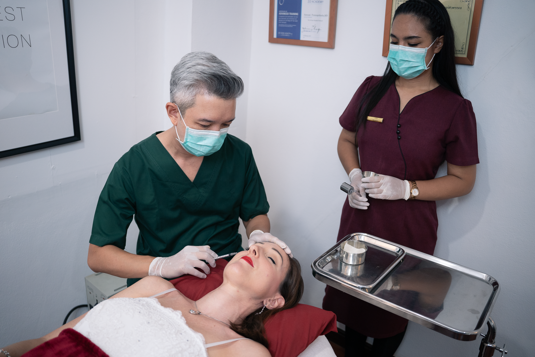 "Ponsarin Clinic doctor in green scrubs performing a botox injection on a client's crowfeet, with a masked assistant preparing supplies nearby."
