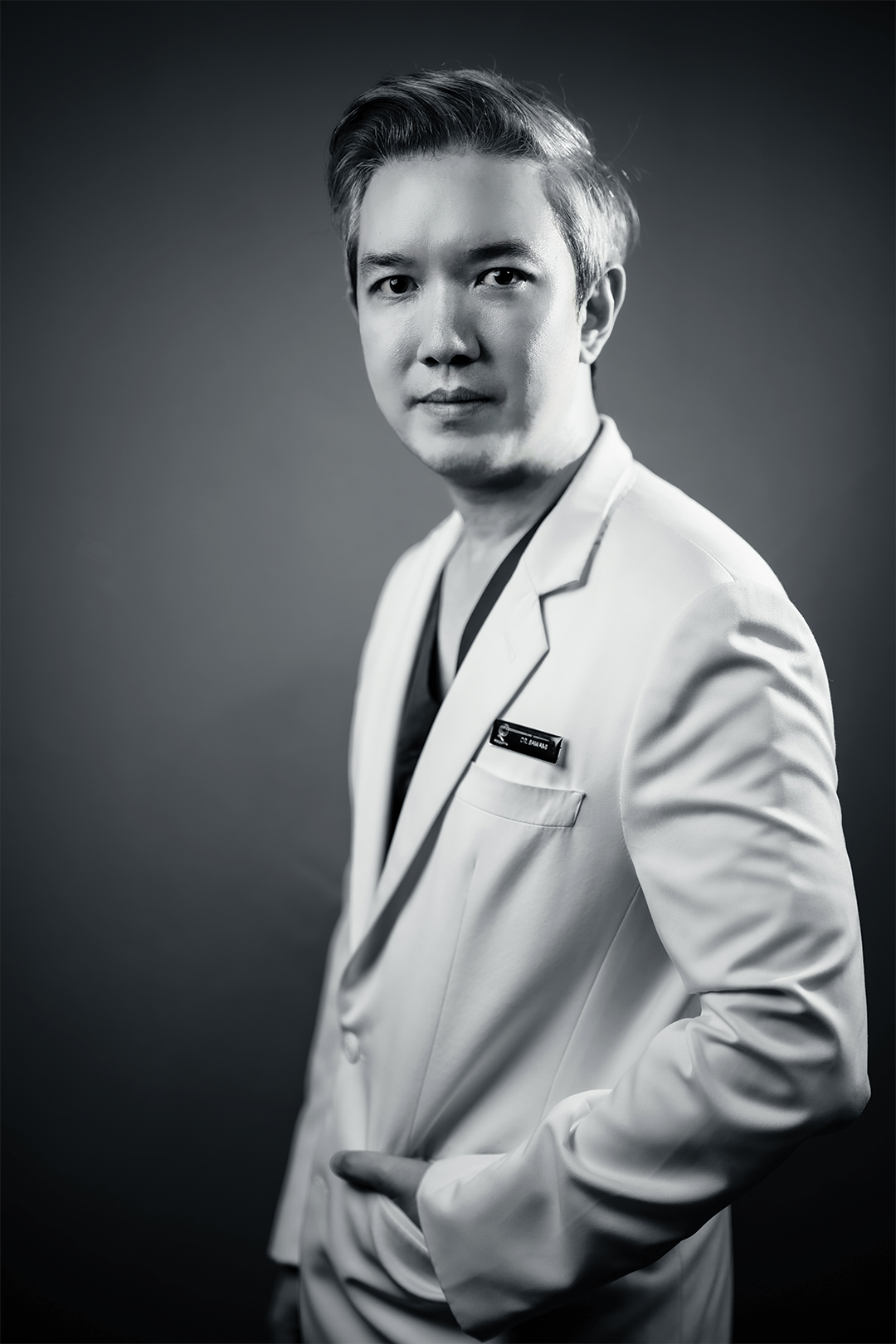 Black and white portrait of Dr.Sawami in a white medical coat with a name tag, looking directly at the camera with a confident expression.