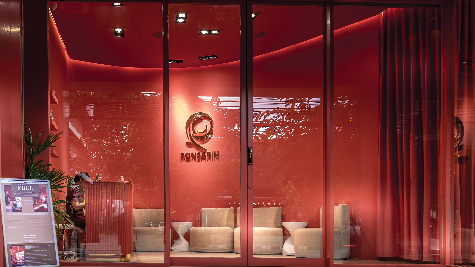 Interior view of a modern, stylish office or lounge with red glass walls, beige chairs, a neon sign logo reading "PONSARIN," and greenery visible through the glass
