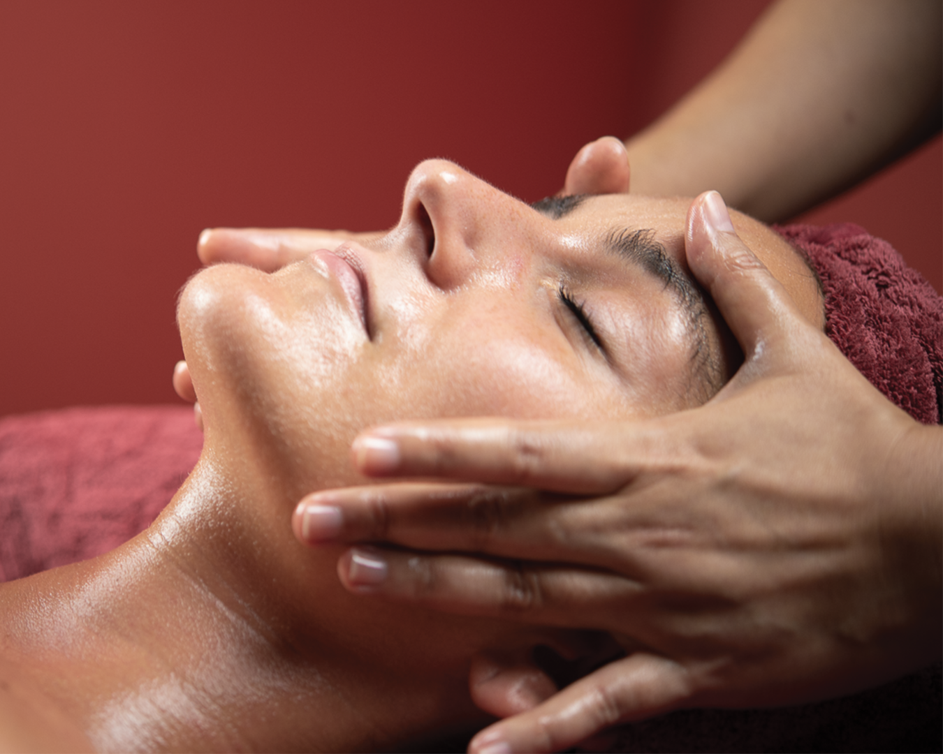 Close-up of a therapist's hands cradling and massaging a client's face during a facial treatment at Ponsarin Aesthetic & Medical Spa, with a burgundy backdrop.