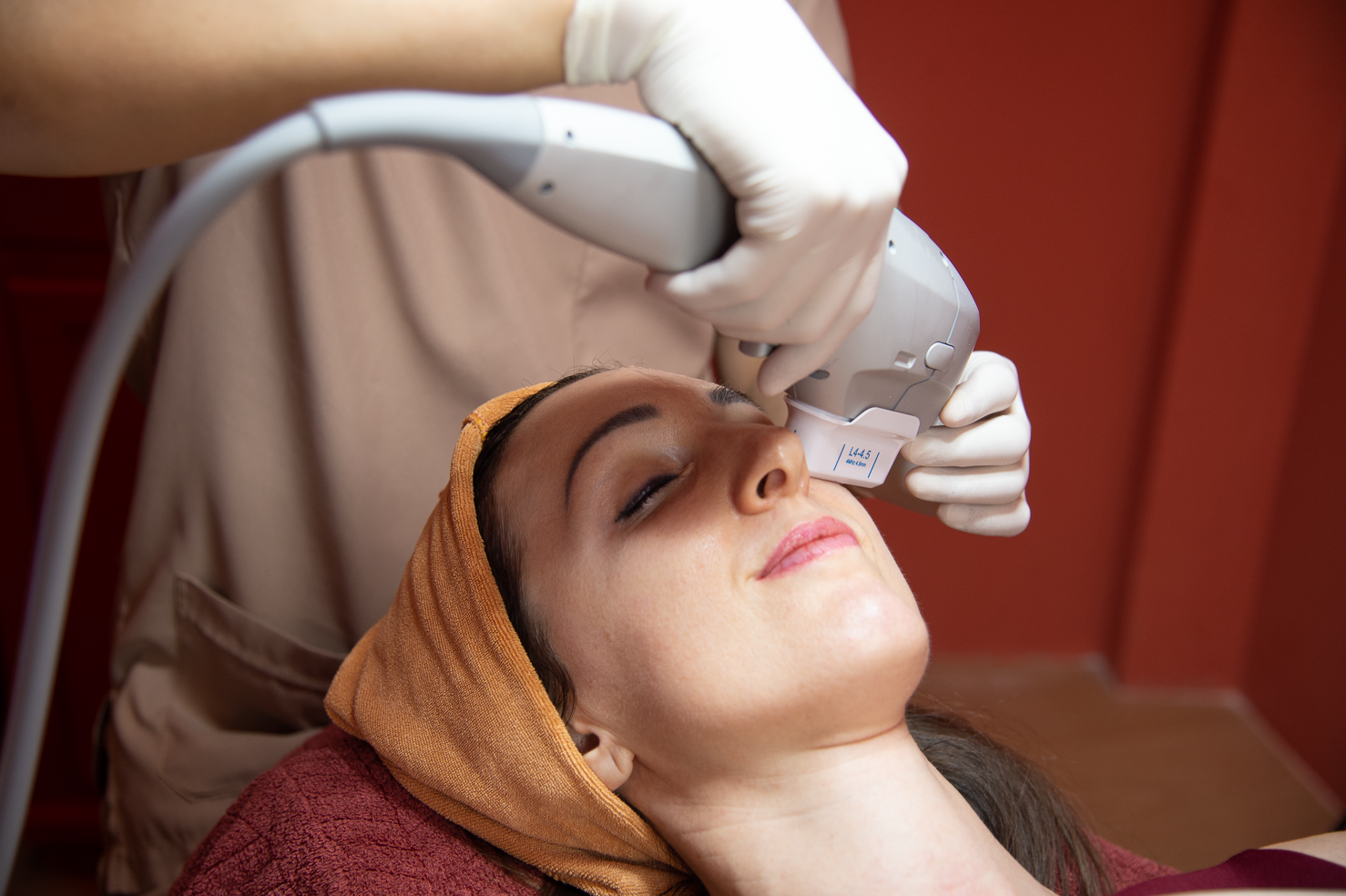 Ponsarin clinic doctor using a Ultra Lift device for a facial treatment on a client wearing a hair towel wrap, close-up under a magnifying lamp.