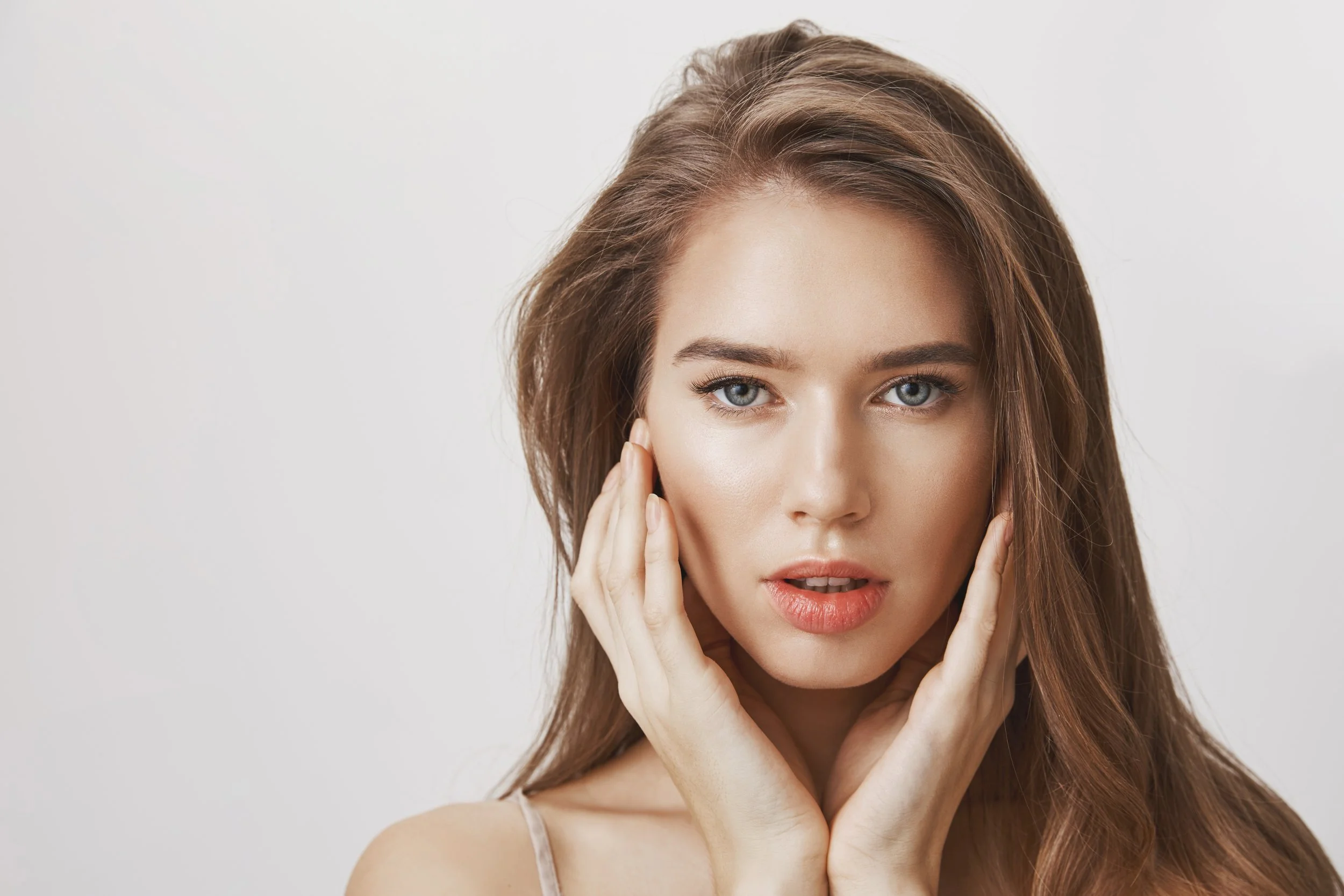 Close-up portrait of a woman with long brown hair and blue eyes, hands framing her face, used to promote dermal filler treatment at Ponsarin Clinic.