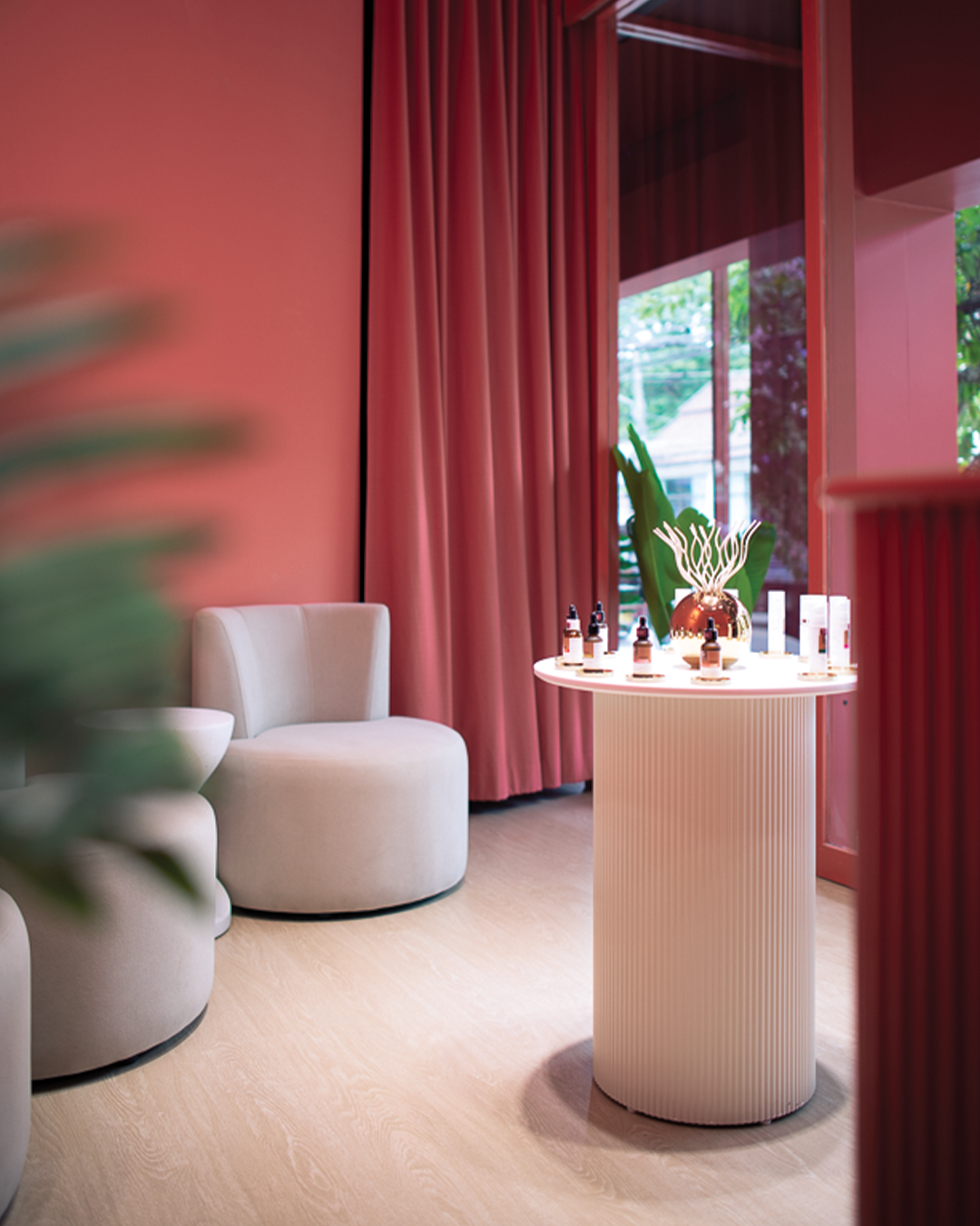 Clinic lounge interior with round white armchairs and ottoman, coral pink curtains, and a white pedestal table displaying skincare product bottles, with tropical foliage and a window in the background.