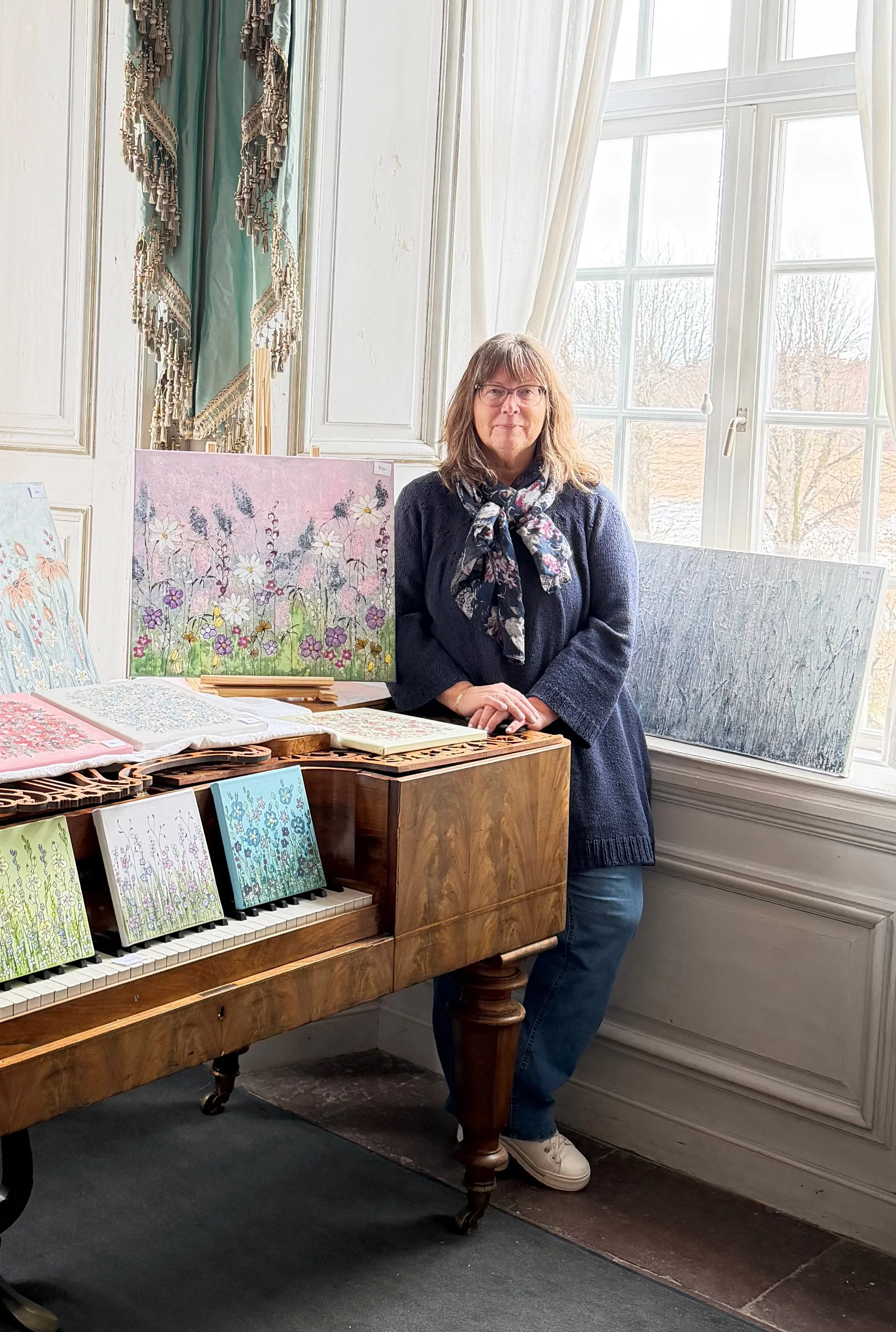 Kvinna står bredvid ett piano omgiven av konstverk med blomstermotiv.