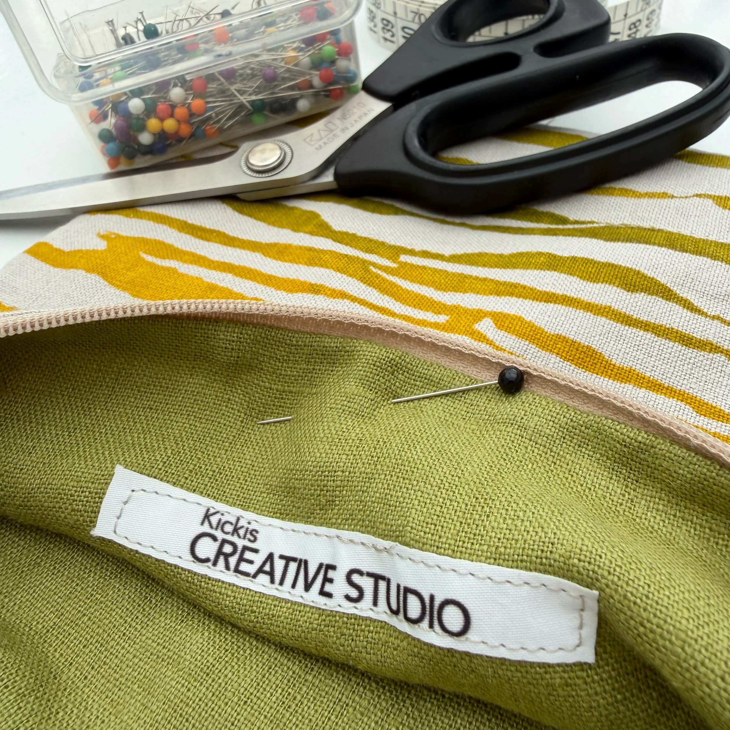 En sax, en öppen plastlåda med handnålar av olika färger och storlekar, och ett lapplapp med texten 'KICKIS CREATIV STUDIO' ligger på ett grönt tyg.