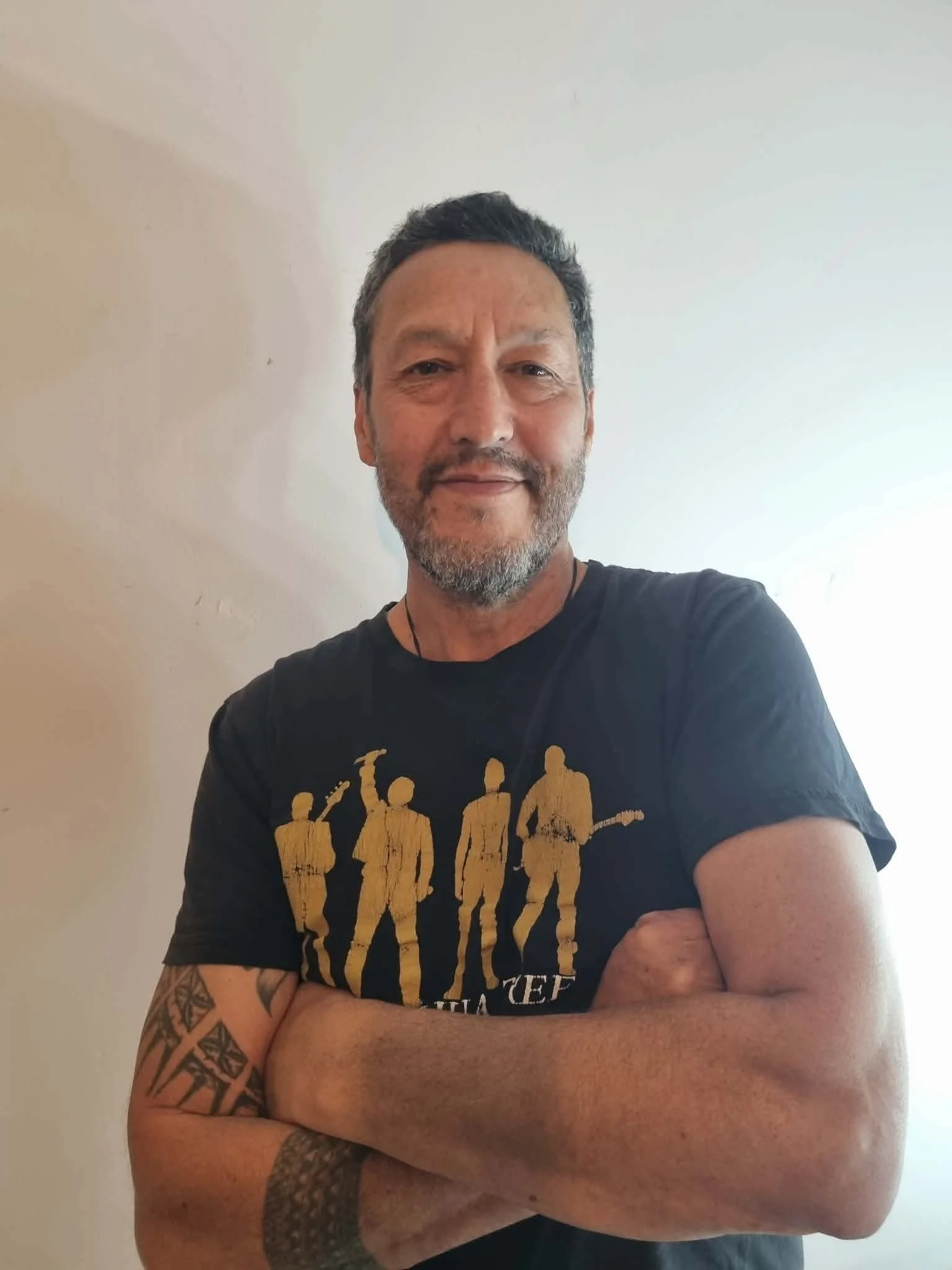 A middle-aged man with short dark hair, a beard, and tattoos on his arms, standing with his arms crossed, wearing a black T-shirt with gold and white graphics, smiling slightly, in front of a plain light-colored wall.