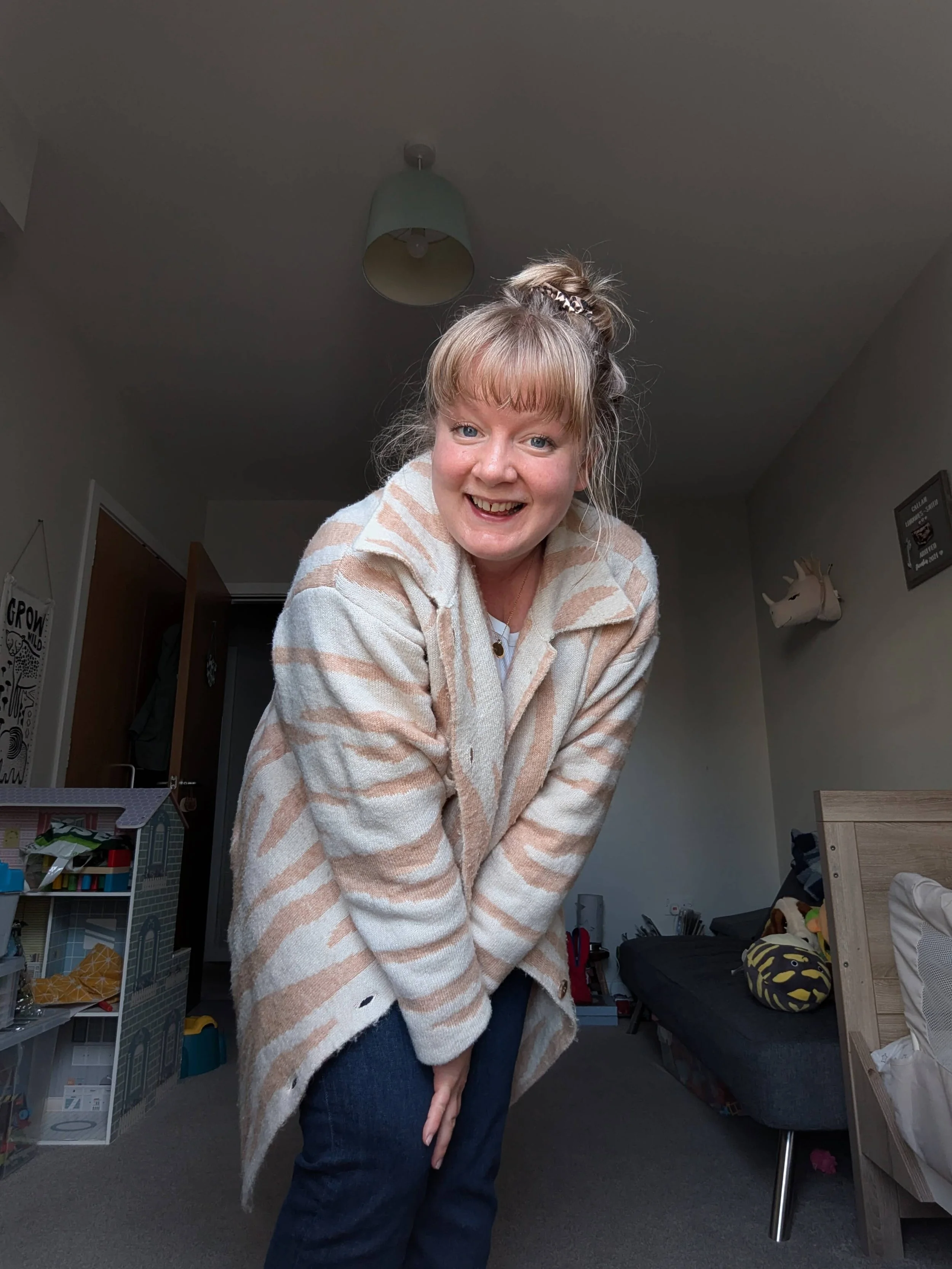 A woman with light hair in a high ponytail, wearing a striped beige and white coat, leaning forward and smiling in a living room.