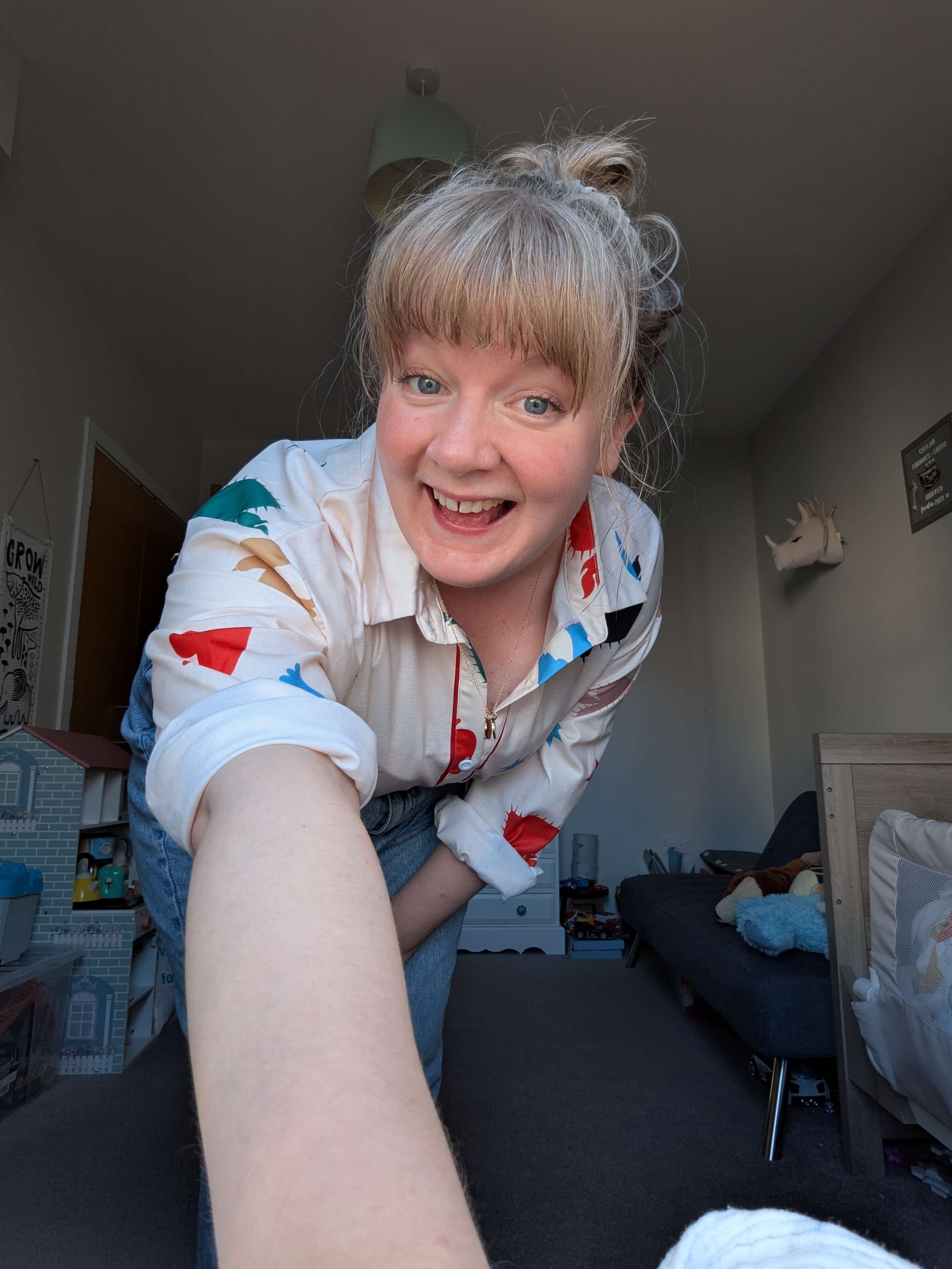 A woman with blonde hair in a messy bun, blue eyes, and a wide smile, leaning toward the camera in a cluttered living room with toys, a sofa, and a unicorn wall decoration.