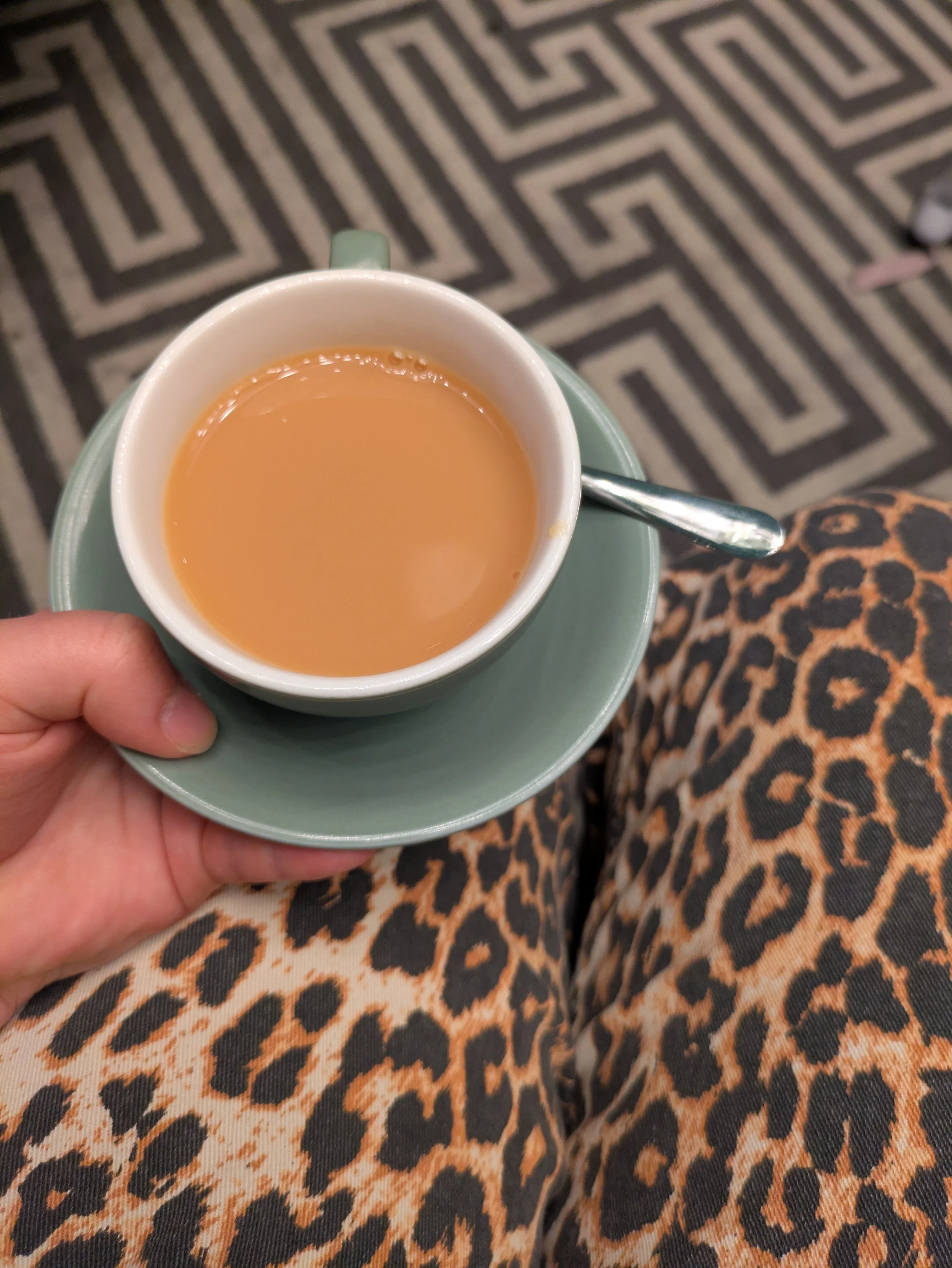 Top view of a hand holding a coffee cup filled with light brown tea, with a silver spoon resting on the saucer. The background features a patterned black and beige carpet, and the person is wearing leopard print pants.
