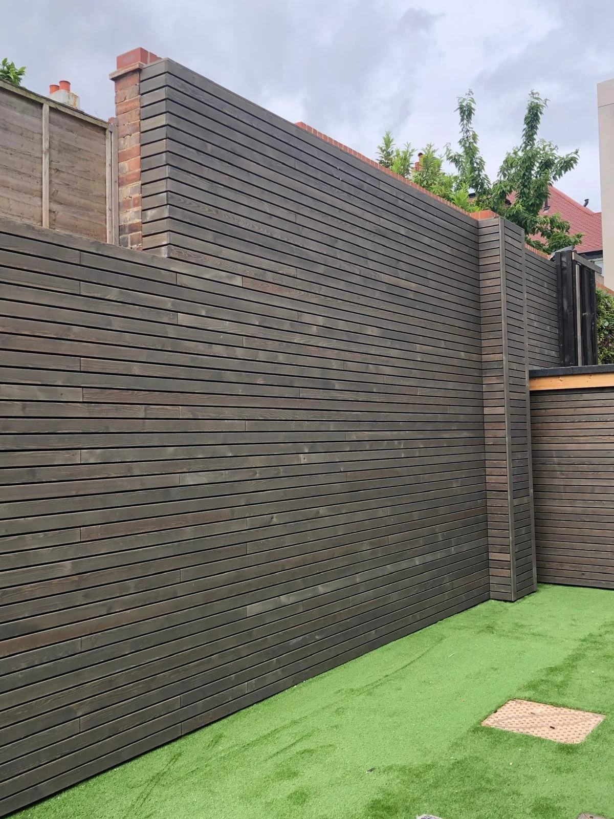 A tall enclosure made of horizontal wooden slats with a small square metal grate on synthetic green grass, with trees and rooftops in the background.