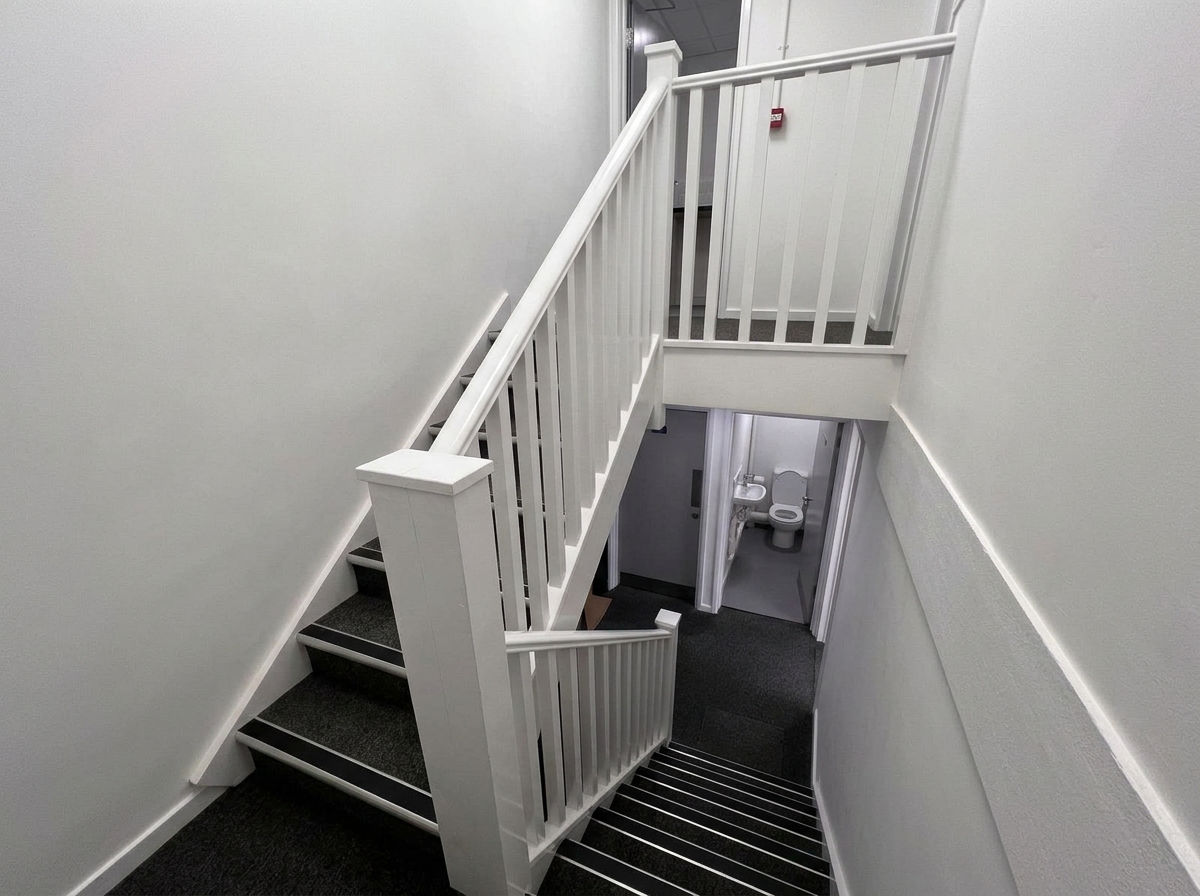 Interior view of a staircase with black carpeted steps and white railing, leading to a lower level with a visible bathroom that has a white toilet and sink.