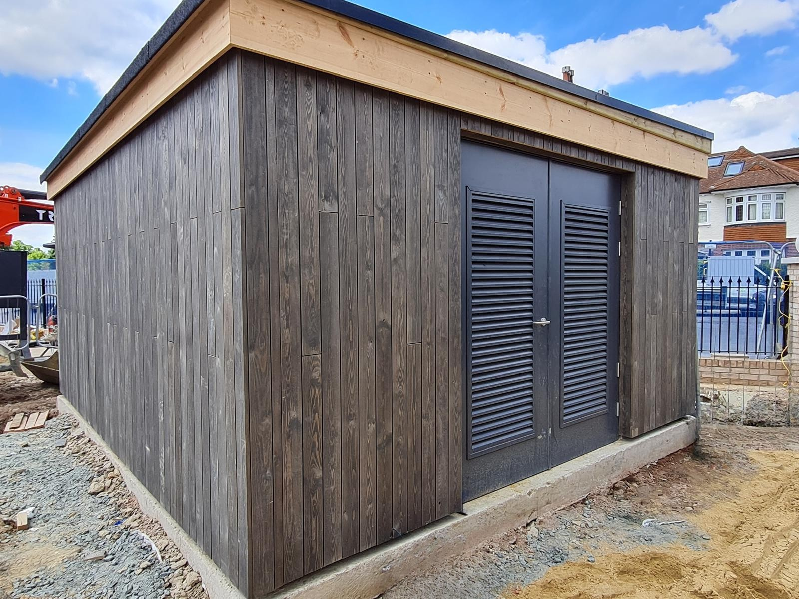Small shed with dark wood exterior and black door with louvered vents, situated on a concrete foundation, under blue sky with clouds.