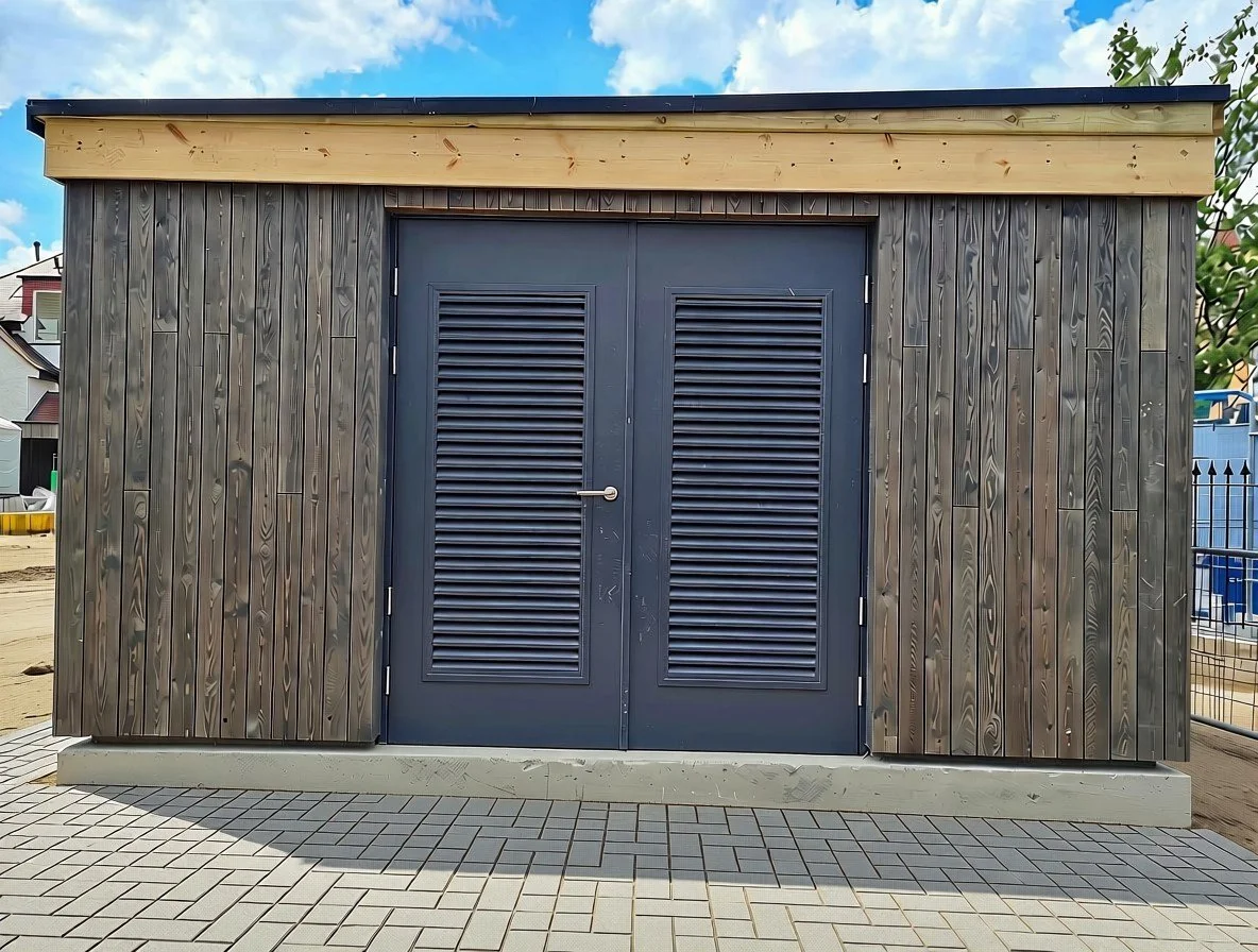 Exterior of a small wooden building with dark gray double doors with louvered panels, surrounded by a brick paver walkway and some greenery on the right side.