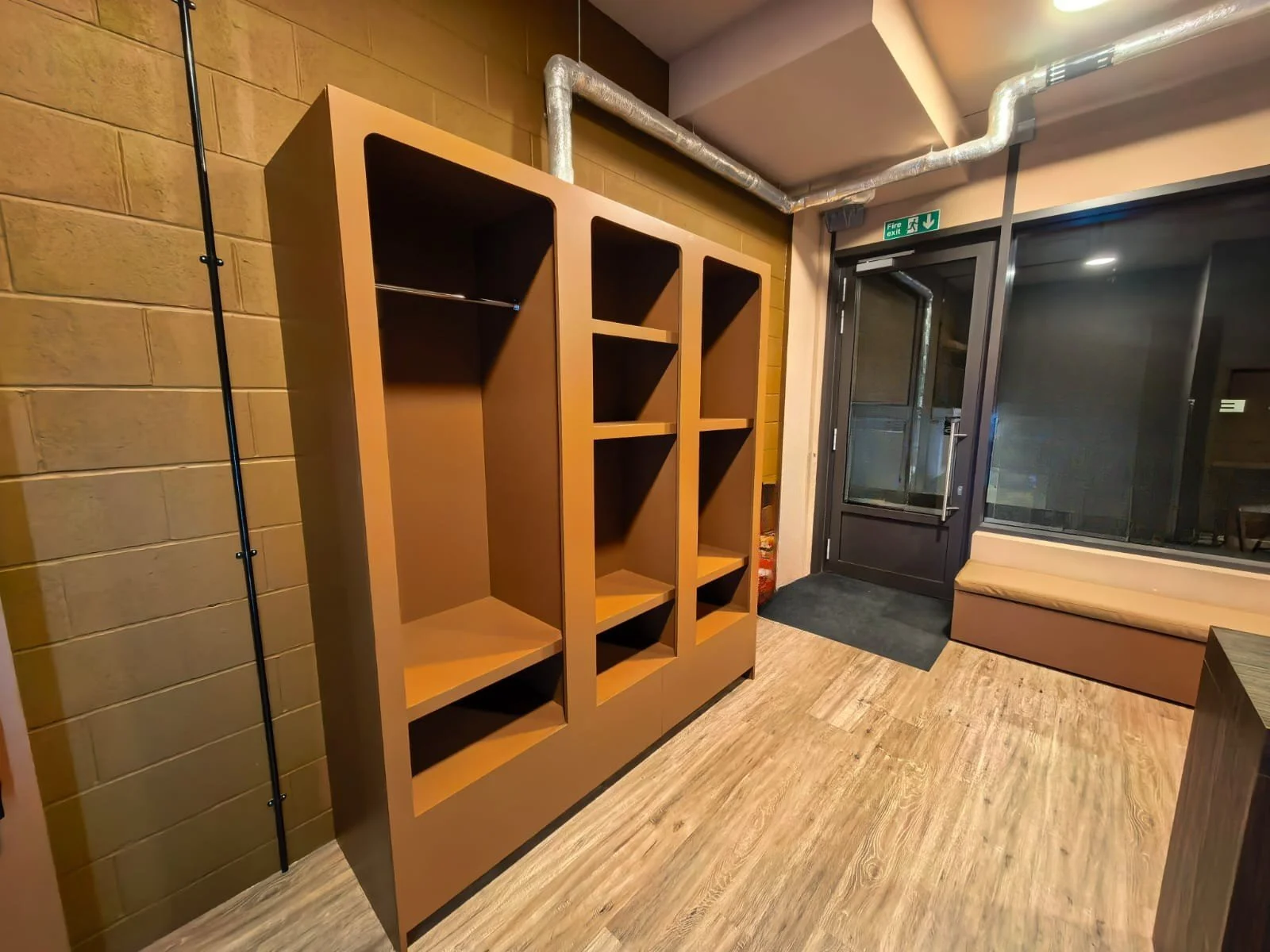 Empty beige wooden bookshelf near a glass door and US 'Fire Exit' sign in a modern indoor space.
