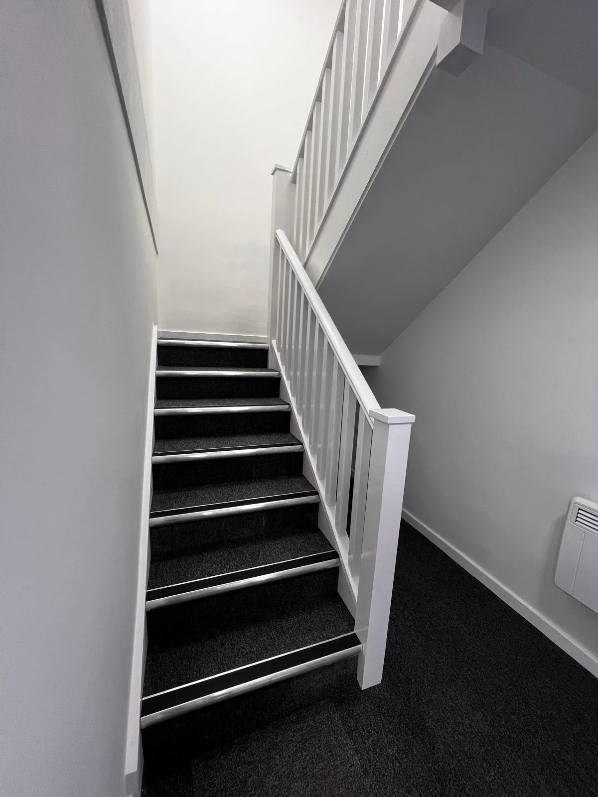 Interior view of a stairway with black carpeted steps, white railing, and grey walls.